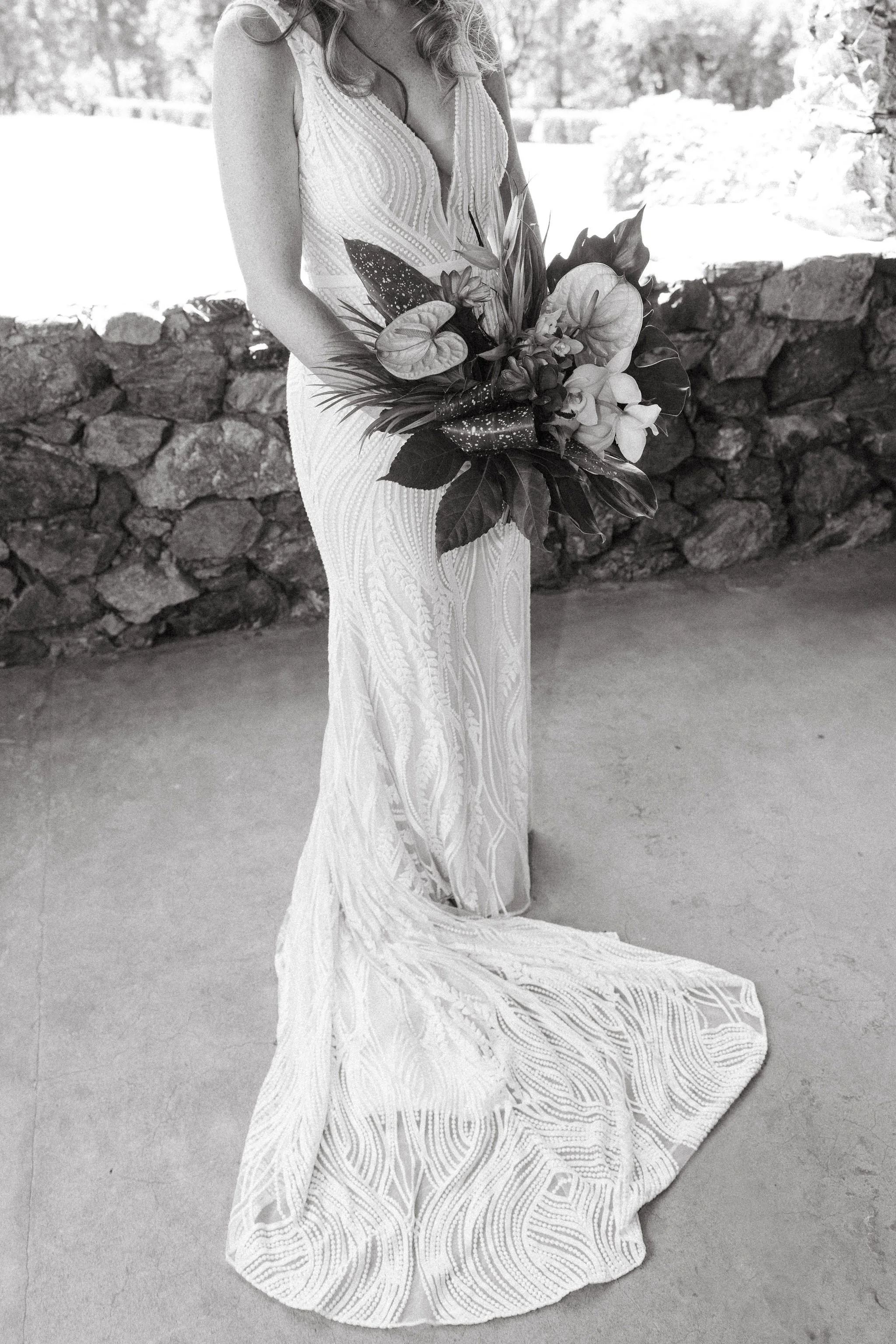 A woman in a white lace dress standing outdoors, holding a large bouquet of flowers.
