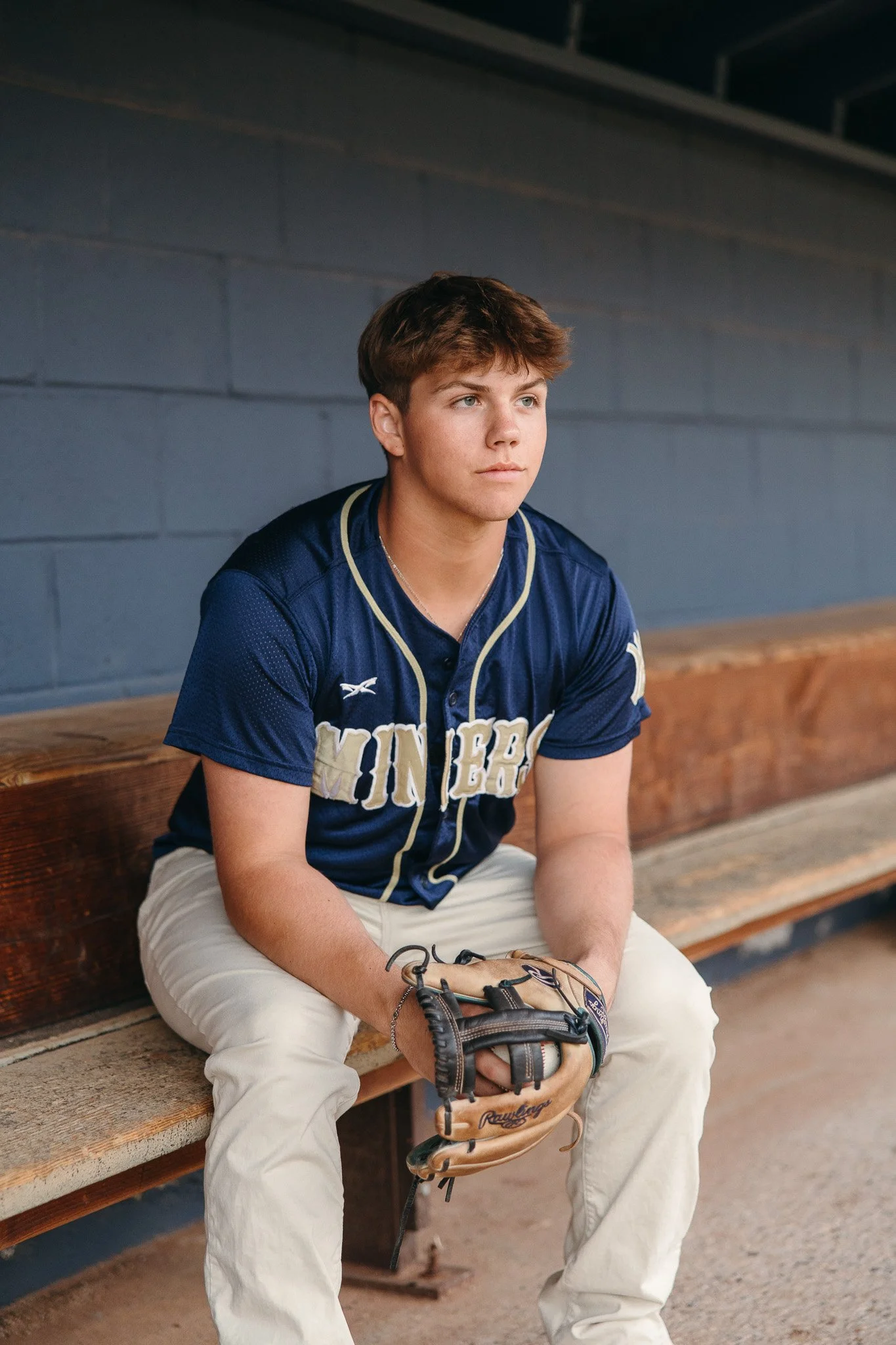 Young man sitting on a wooden bench in a baseball dugout, wearing a navy blue baseball jersey with white lettering and white pants, holding a baseball glove. He has short brown hair and is looking off to the side with a serious expression.