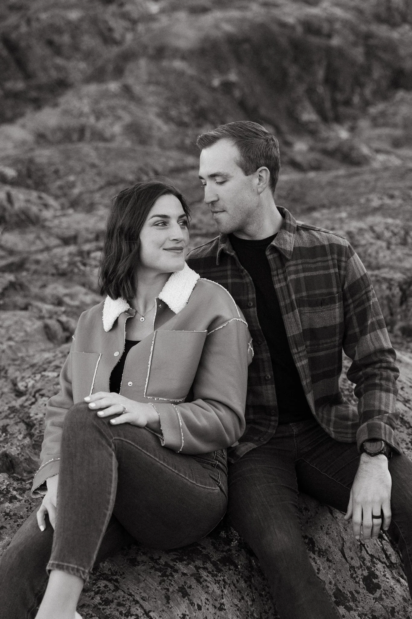 A black-and-white photo of a couple sitting outdoors on a large rock. The woman has shoulder-length hair and is wearing a jacket with a fur collar, while the man has short hair and is wearing a plaid shirt. They are looking at each other warmly, with