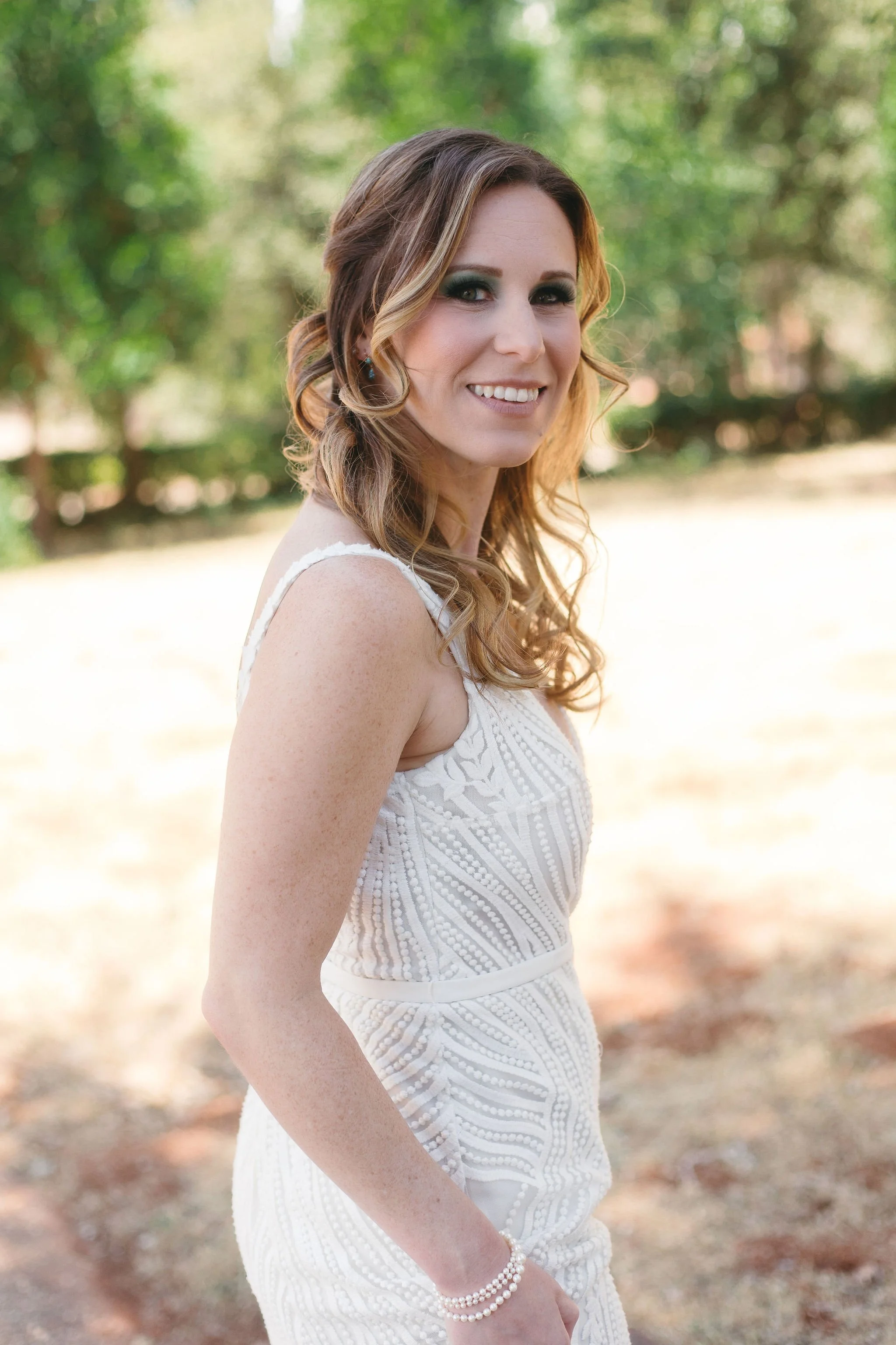 A woman with wavy, shoulder-length hair, wearing a white, sleeveless dress with lace details. She is smiling and standing outdoors with trees in the background, and appears to be at a wedding or special event.
