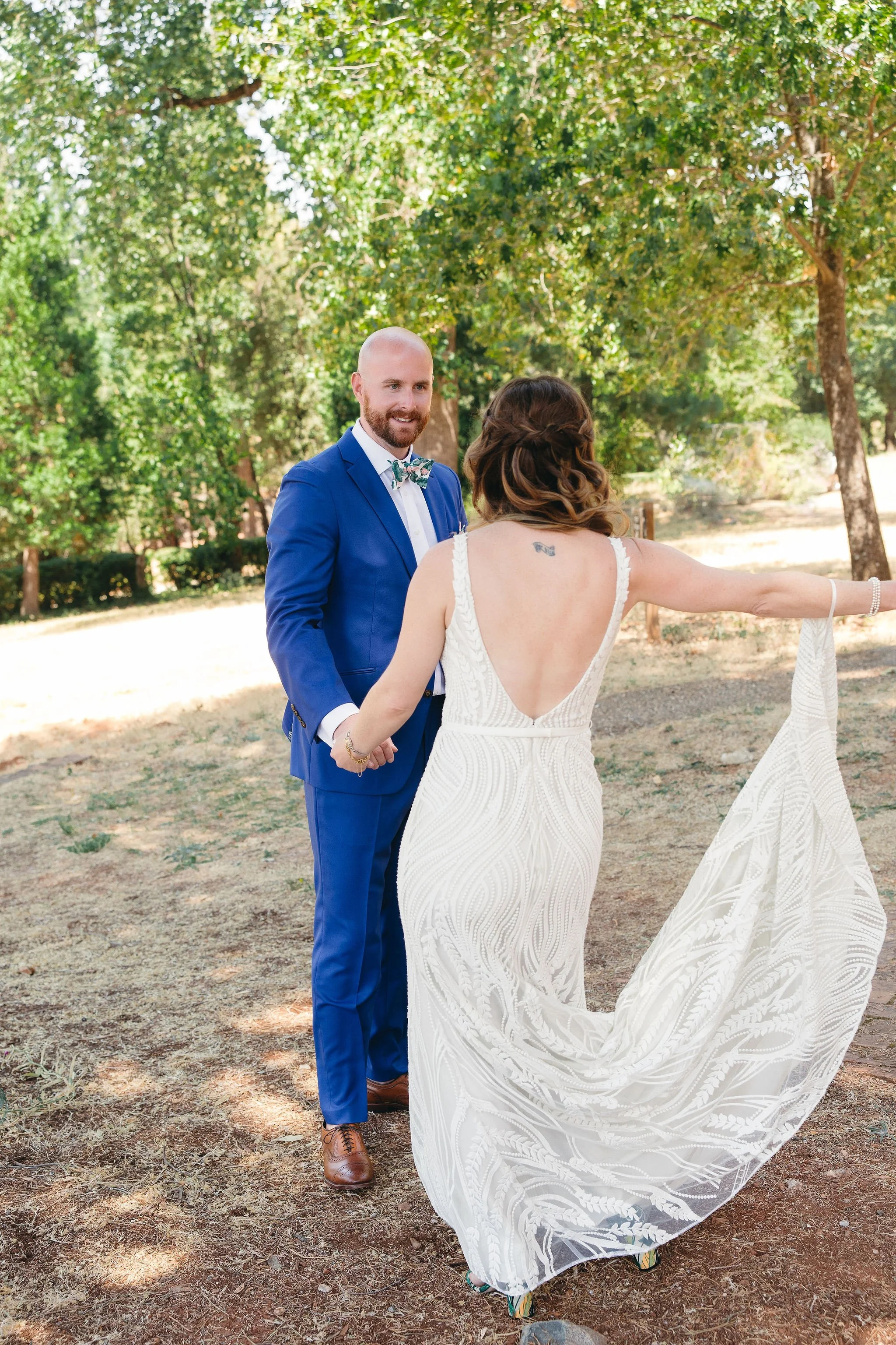A couple holding hands outdoors during a wedding ceremony, with a woman in a white dress and a man in a blue suit, standing under trees on a sunny day.