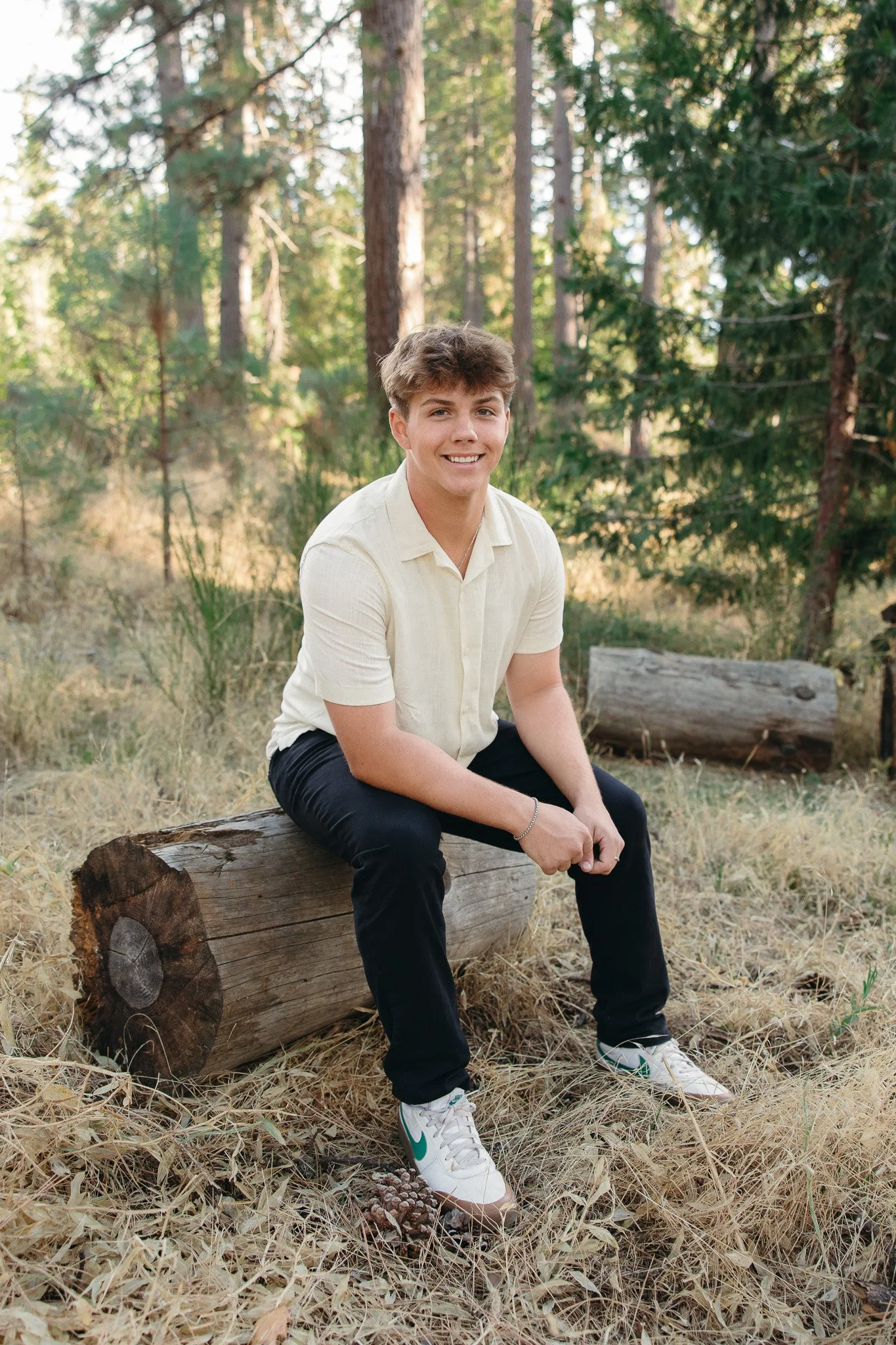 Young man sitting on a tree trunk in a wooded area, smiling at the camera, with tall trees and grass in the background.