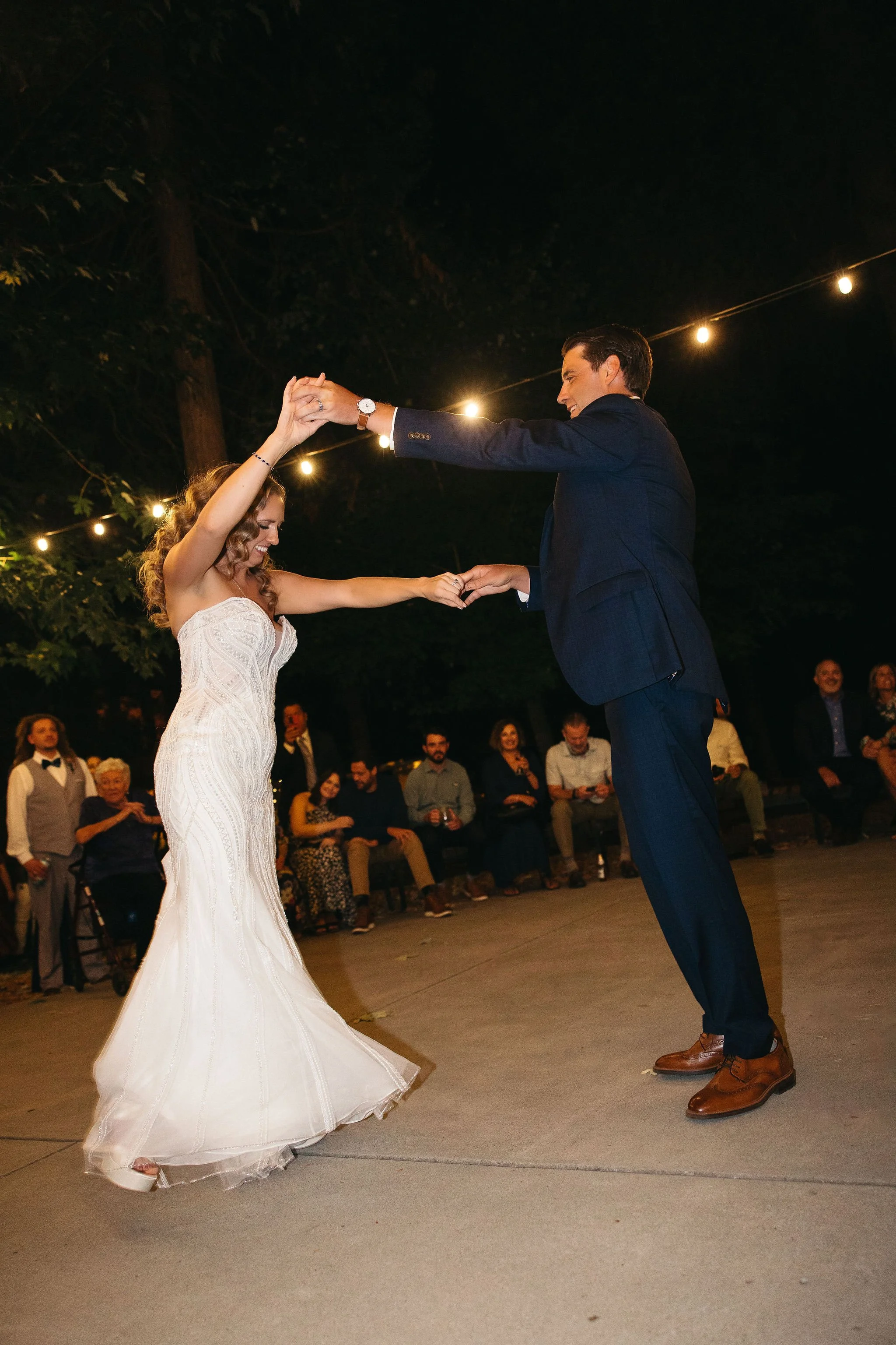 A bride and groom dancing at their wedding reception outdoors at night, with guests sitting on benches in the background and string lights overhead.