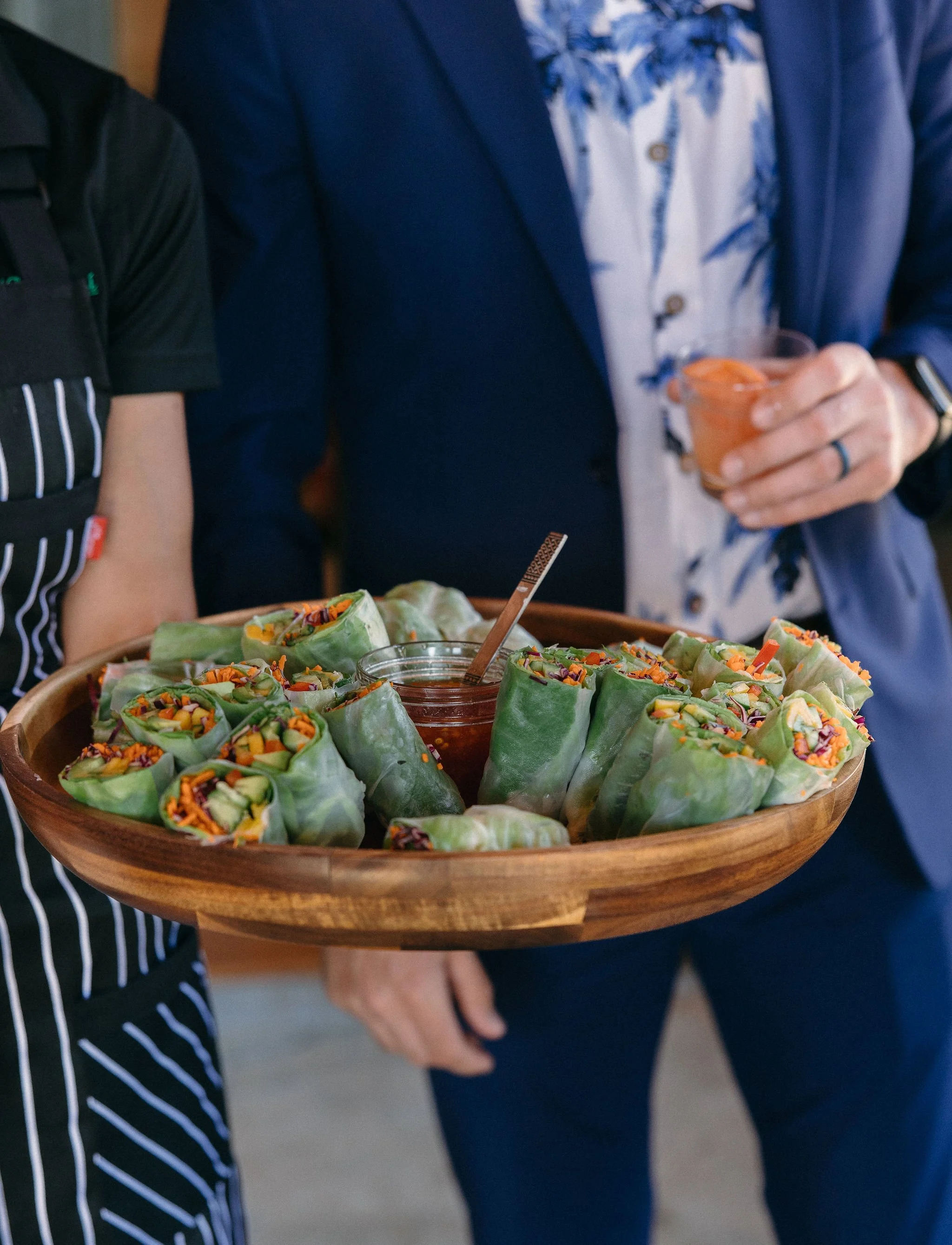A person holding a wooden tray filled with fresh spring rolls with colorful vegetables and a small bowl of dipping sauce in the center, at a social gathering.