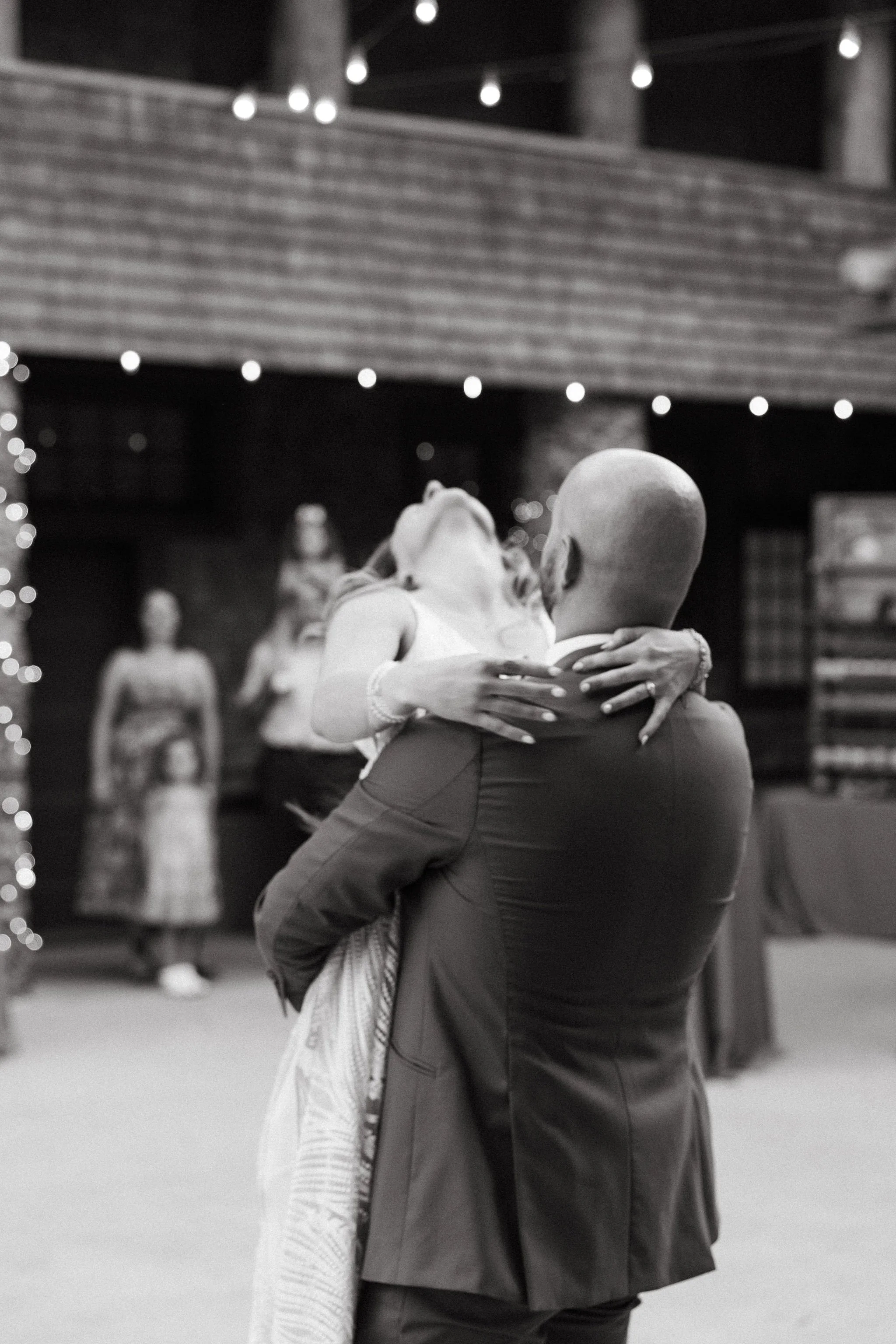 A couple dancing closely, with the woman leaning back and the man holding her, in a dimly lit indoor venue decorated with string lights, with several onlookers in the background.