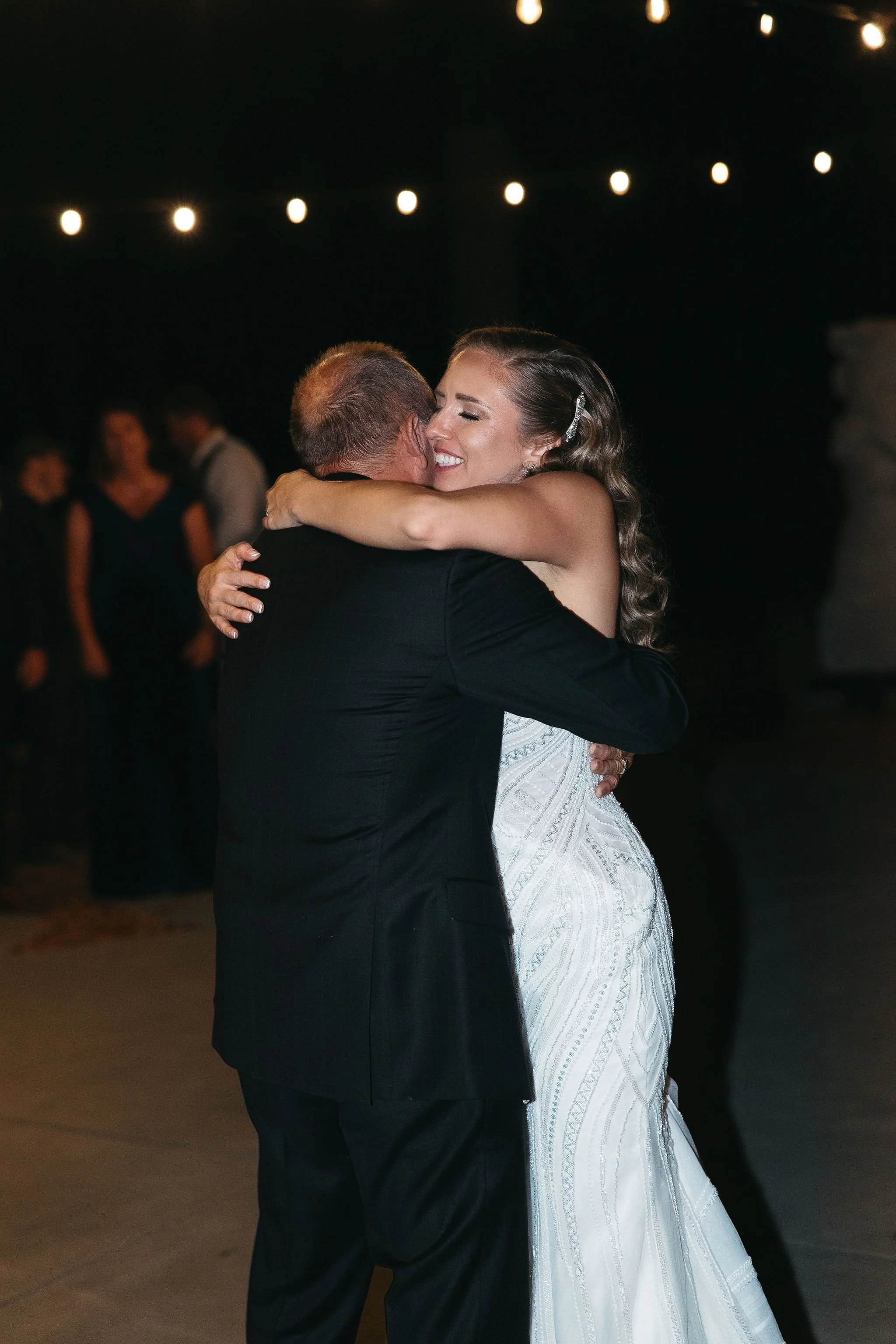 A bride and an older man, possibly her father, are hugging and dancing at a wedding reception. The bride is smiling and wearing a white wedding gown, while the older man is dressed in a black suit. Background includes string lights and a few guests.