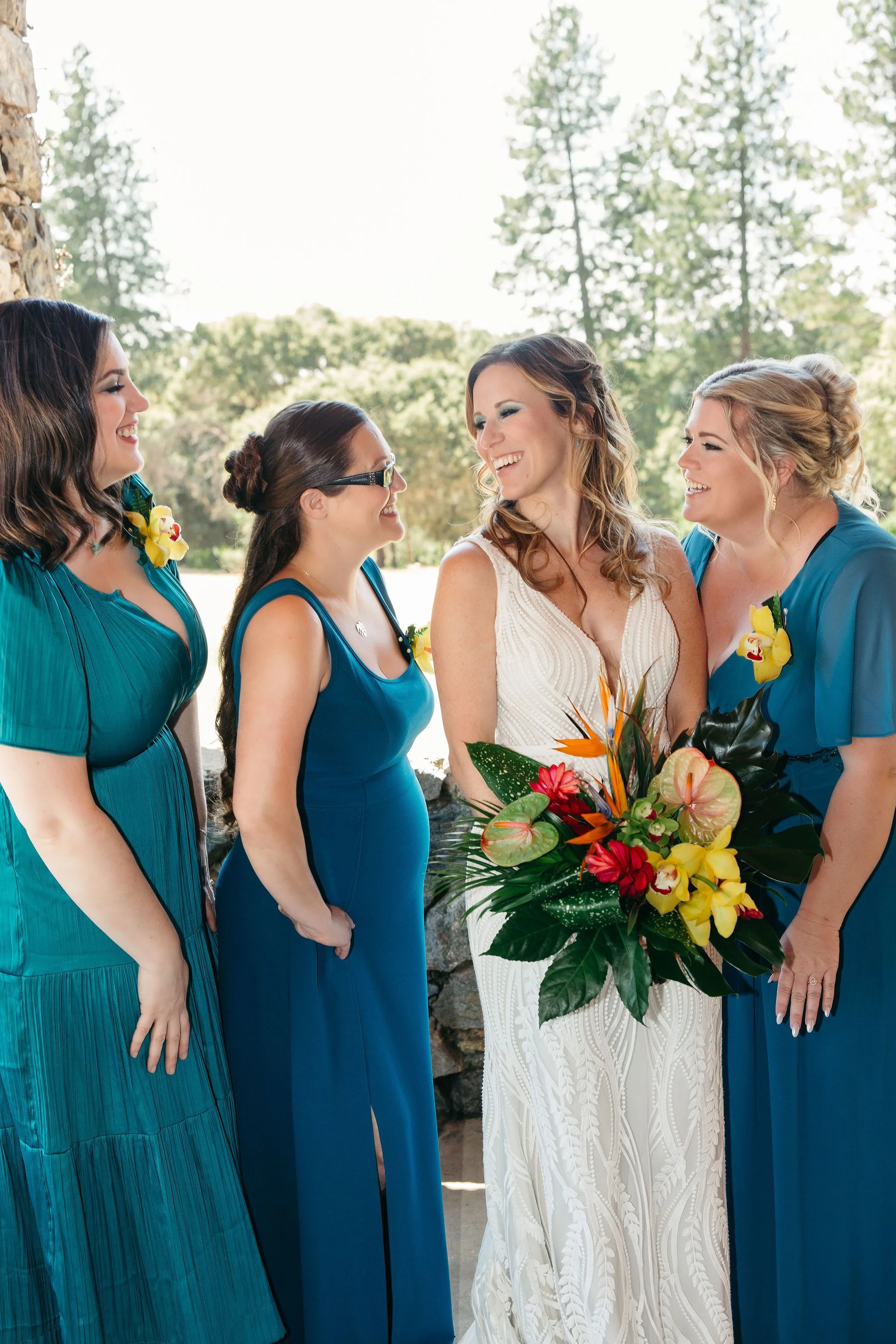 Four women at a wedding, with the bride holding a colorful bouquet, smiling and standing outdoors with trees in the background.