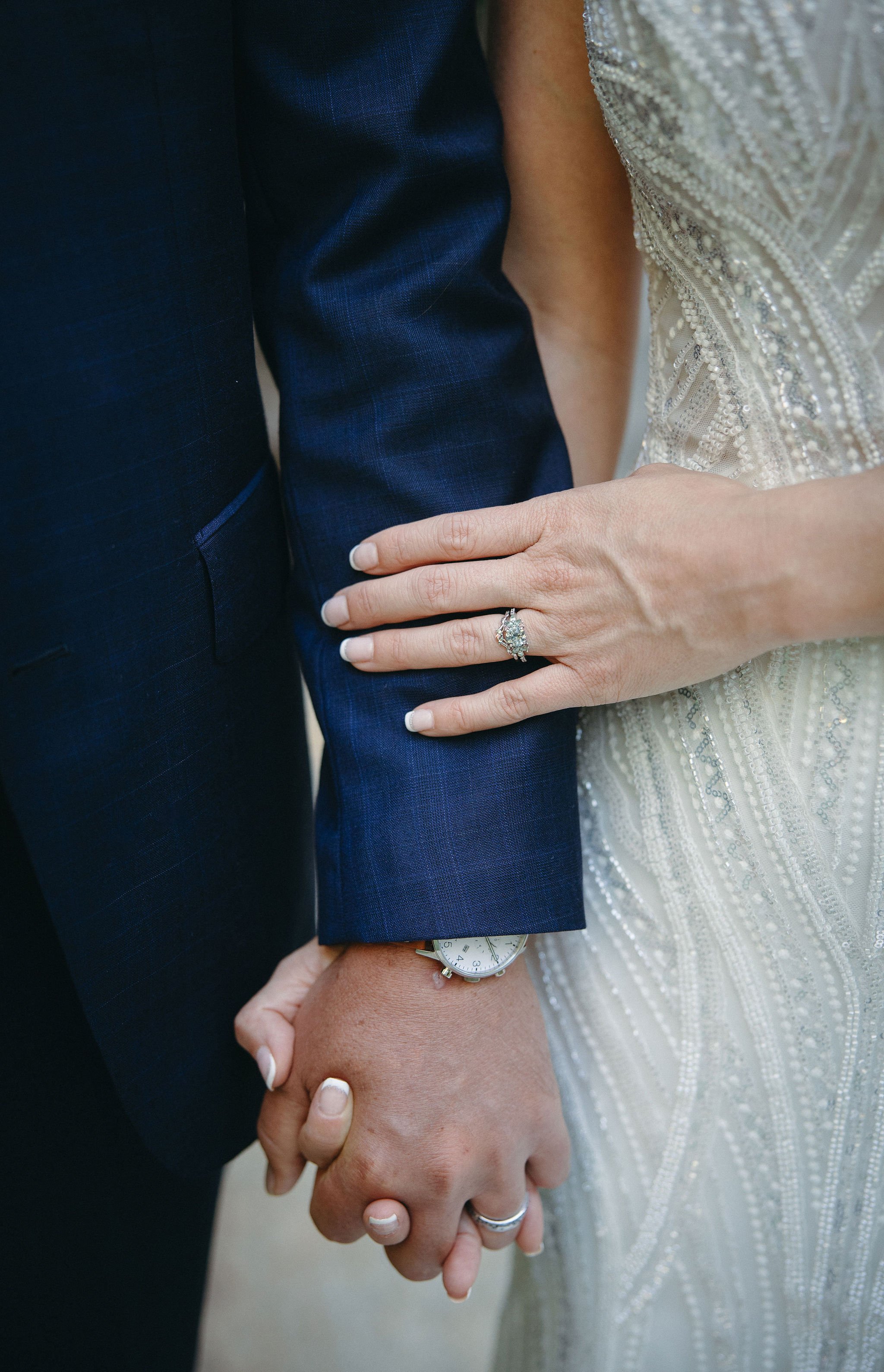 Close-up of a couple holding hands on their wedding day, with a focus on the bride's ring, the groom's watch, and their hands intertwined, showing the bride's elegant dress and the groom's dark suit.