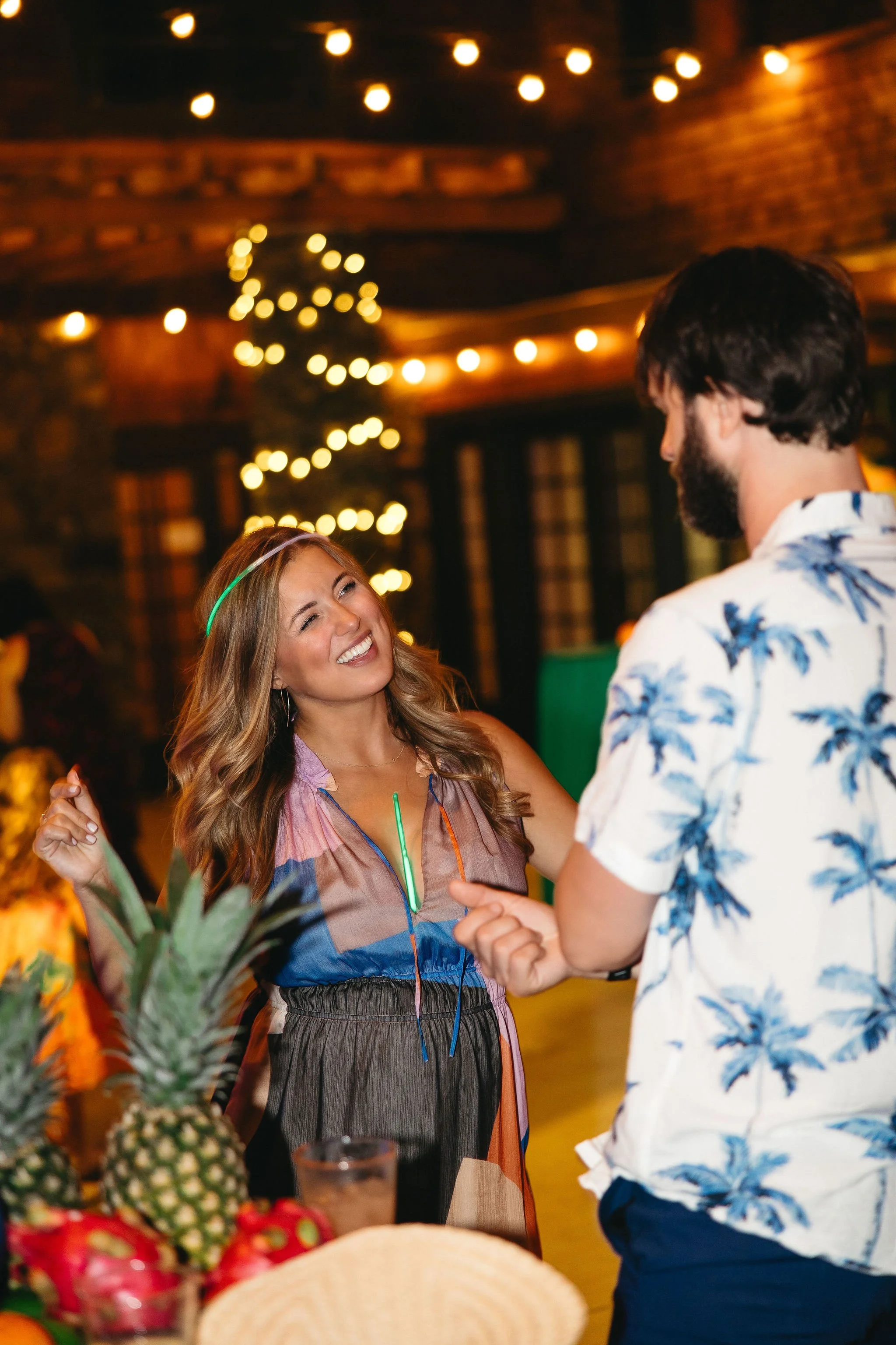 A woman and a man dancing at a party with tropical decorations and string lights.