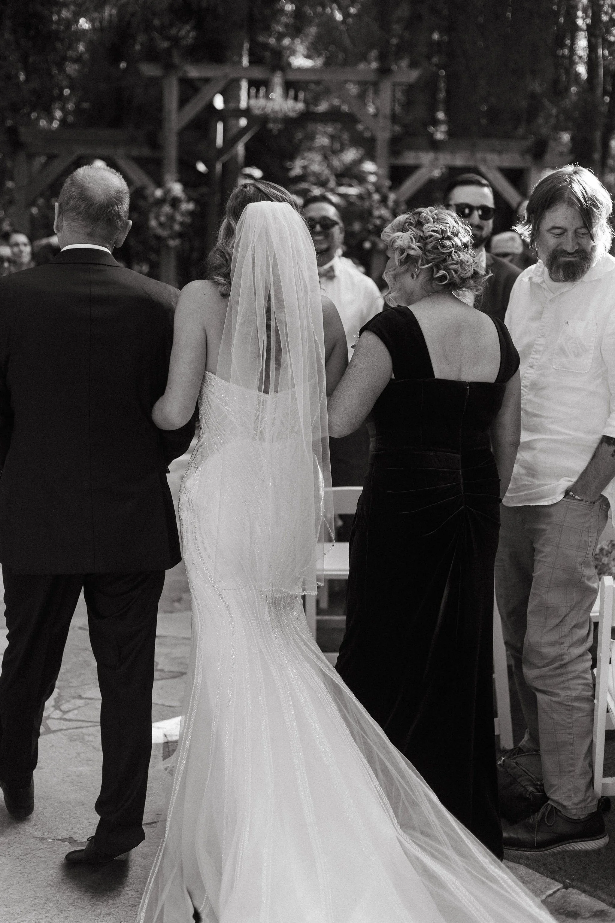 A bride in a wedding dress and veil, accompanied by a man in a suit, standing outdoors at a wedding ceremony surrounded by guests, some of whom are smiling and dressed casually.