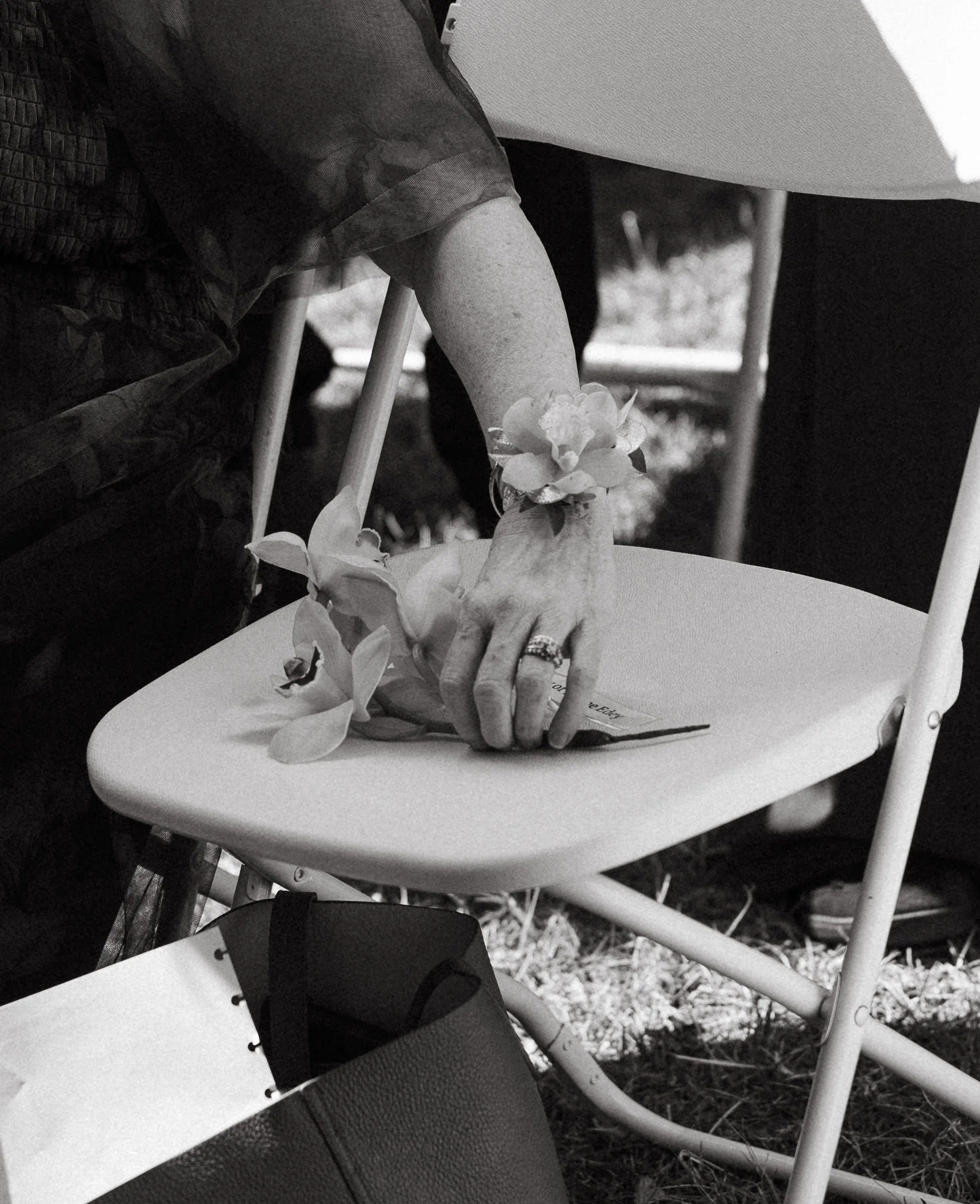 A person sitting at a table, with their hand resting on a chair, wearing a flower bracelet on their wrist and a ring on their finger, holding a flower or small bouquet. The image is in black and white.