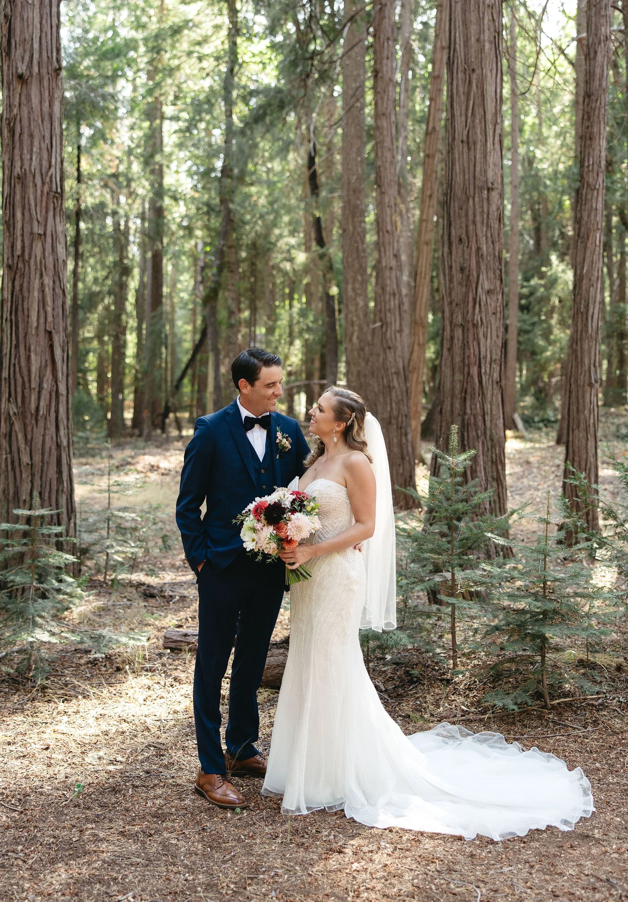 A bride and groom standing together in a forest, gazing at each other, with the bride holding a bouquet of flowers.
