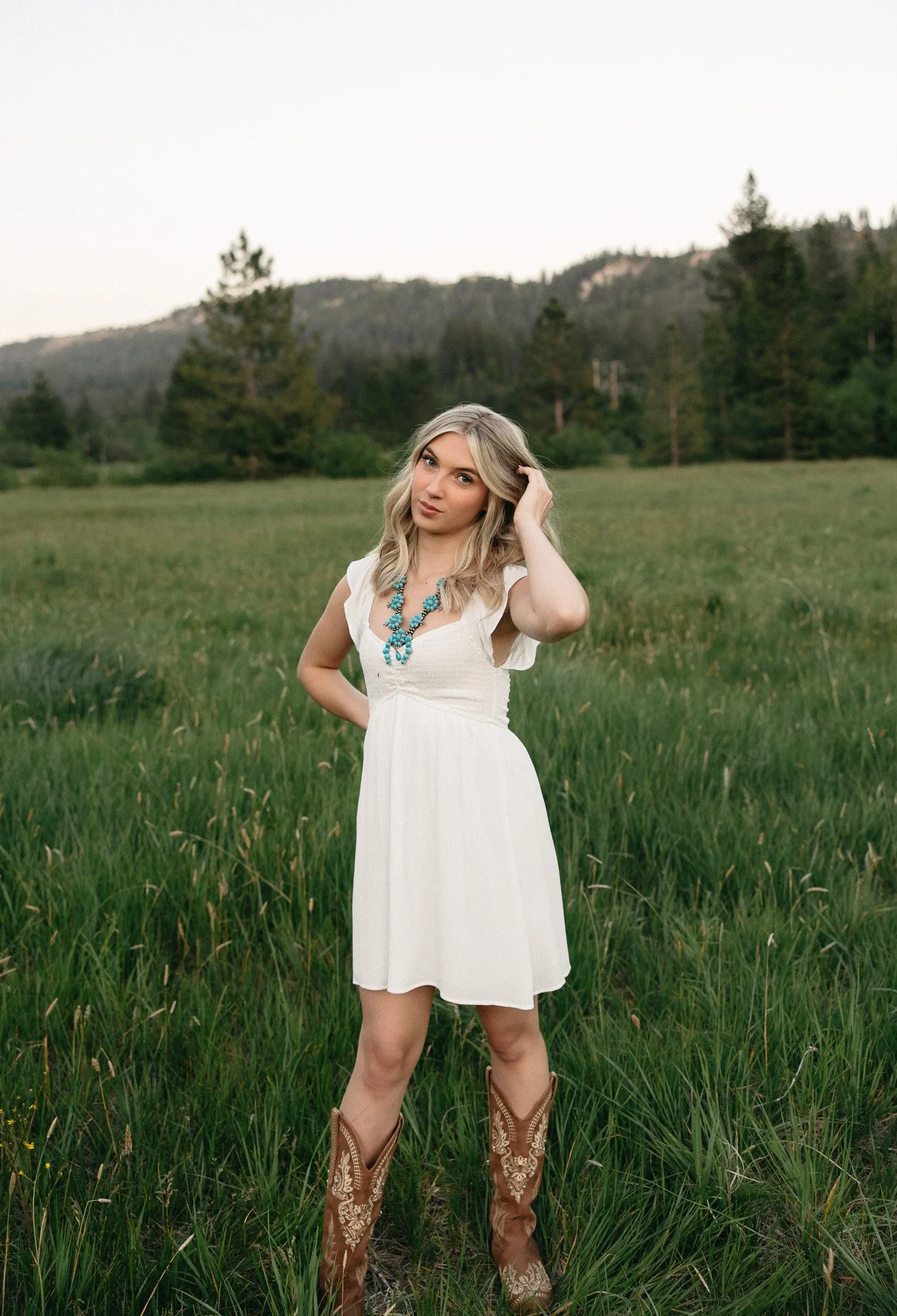 A woman in a white dress and cowboy boots standing in a grassy field with trees and hills in the background.