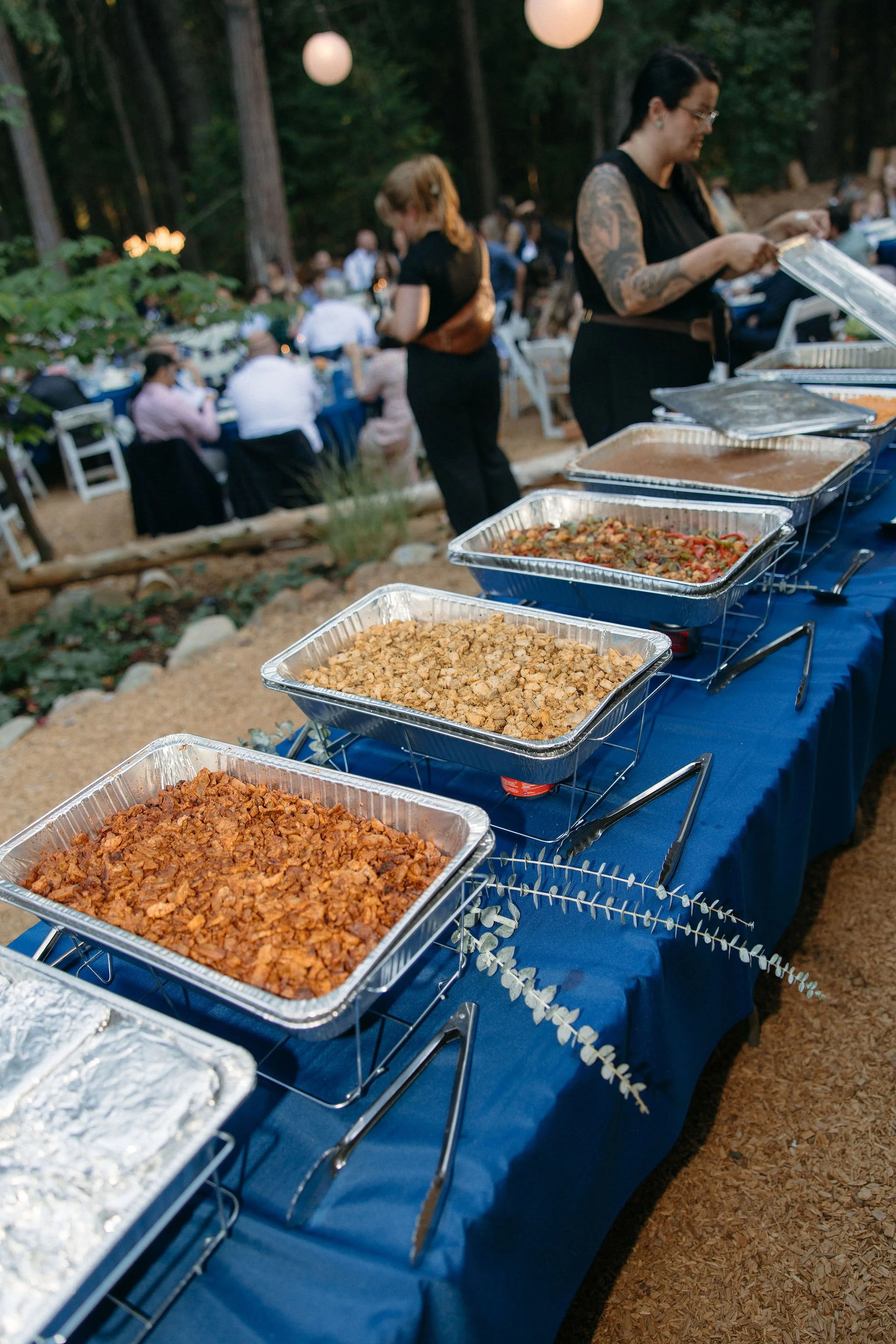 Buffet table with several trays of food at an outdoor event, with people dining in the background in a wooded area