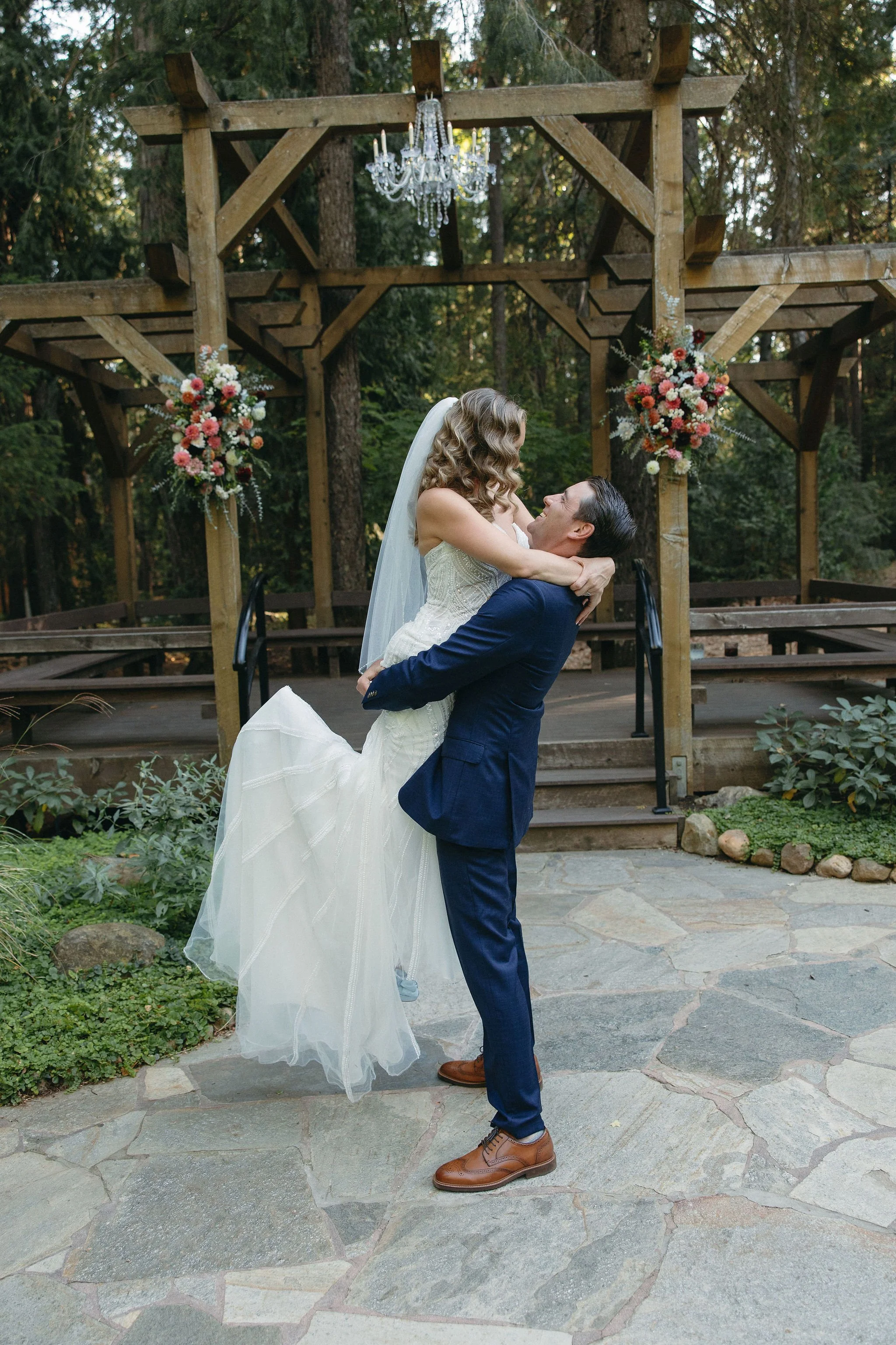 A bride and groom embrace outdoors on a stone patio with wooden stage and floral decorations, forest in the background, during their wedding.