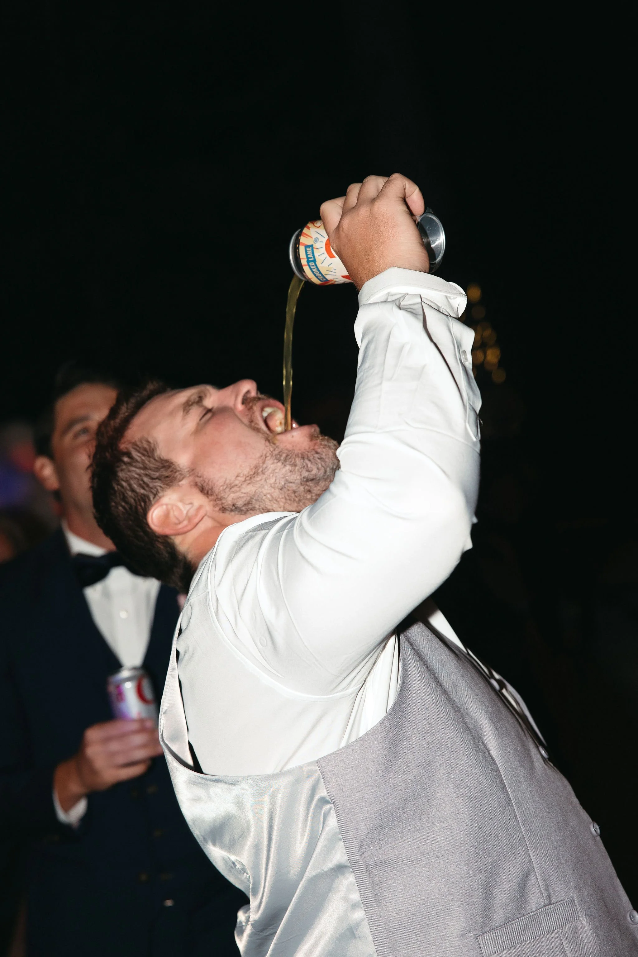 Man in tuxedo and vest pouring a drink into his mouth at a nighttime event.