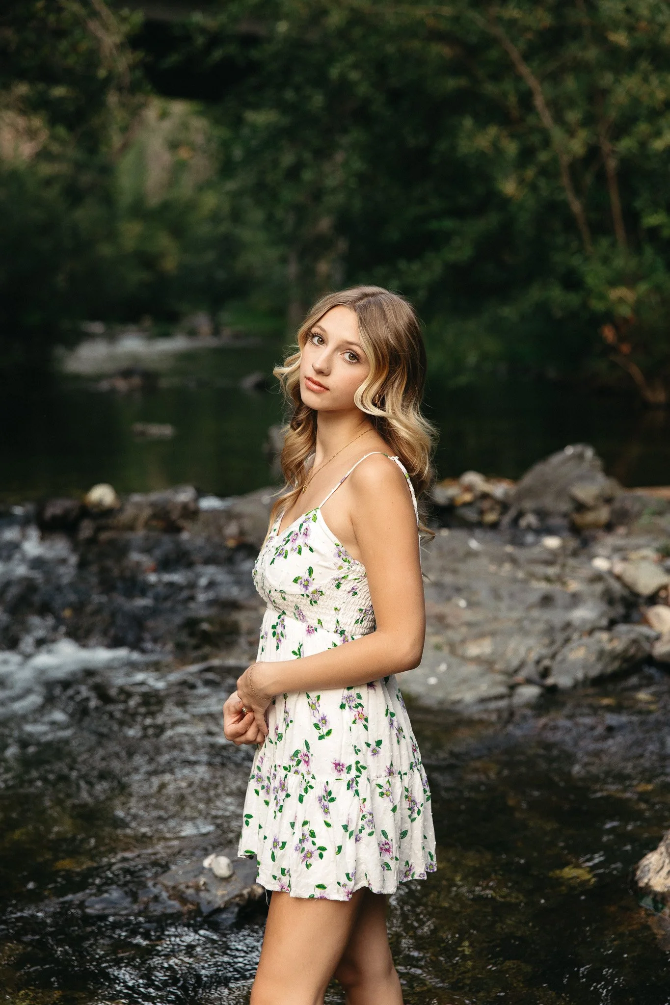 A young woman with wavy blonde hair and light skin standing by a river with rocks and trees in the background, wearing a white dress with purple and green floral pattern.