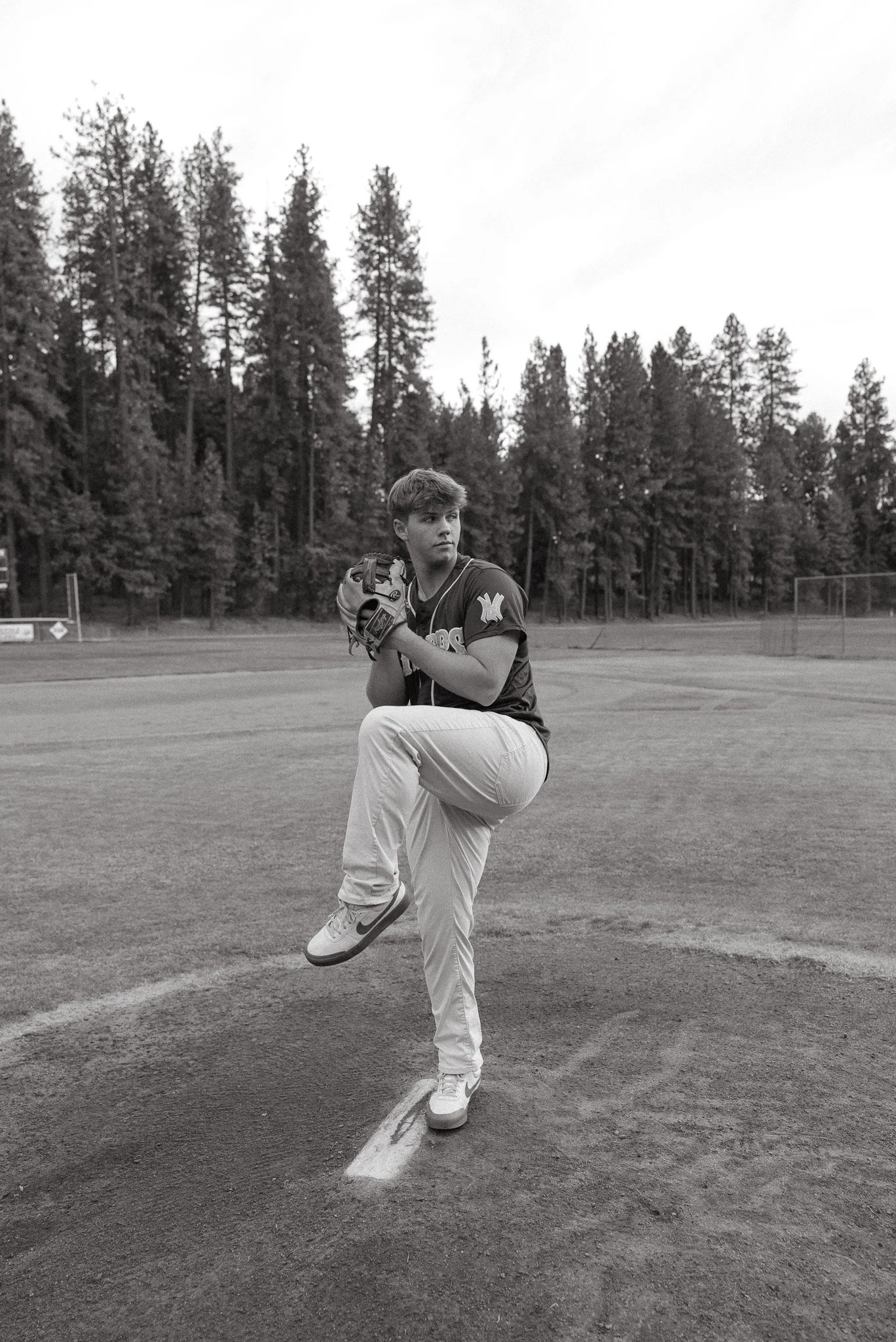 A young male baseball player in uniform is practicing on a baseball field. He is standing on the pitcher's mound, holding a baseball in his glove, with one leg lifted. The field is surrounded by tall trees, and the sky is overcast.