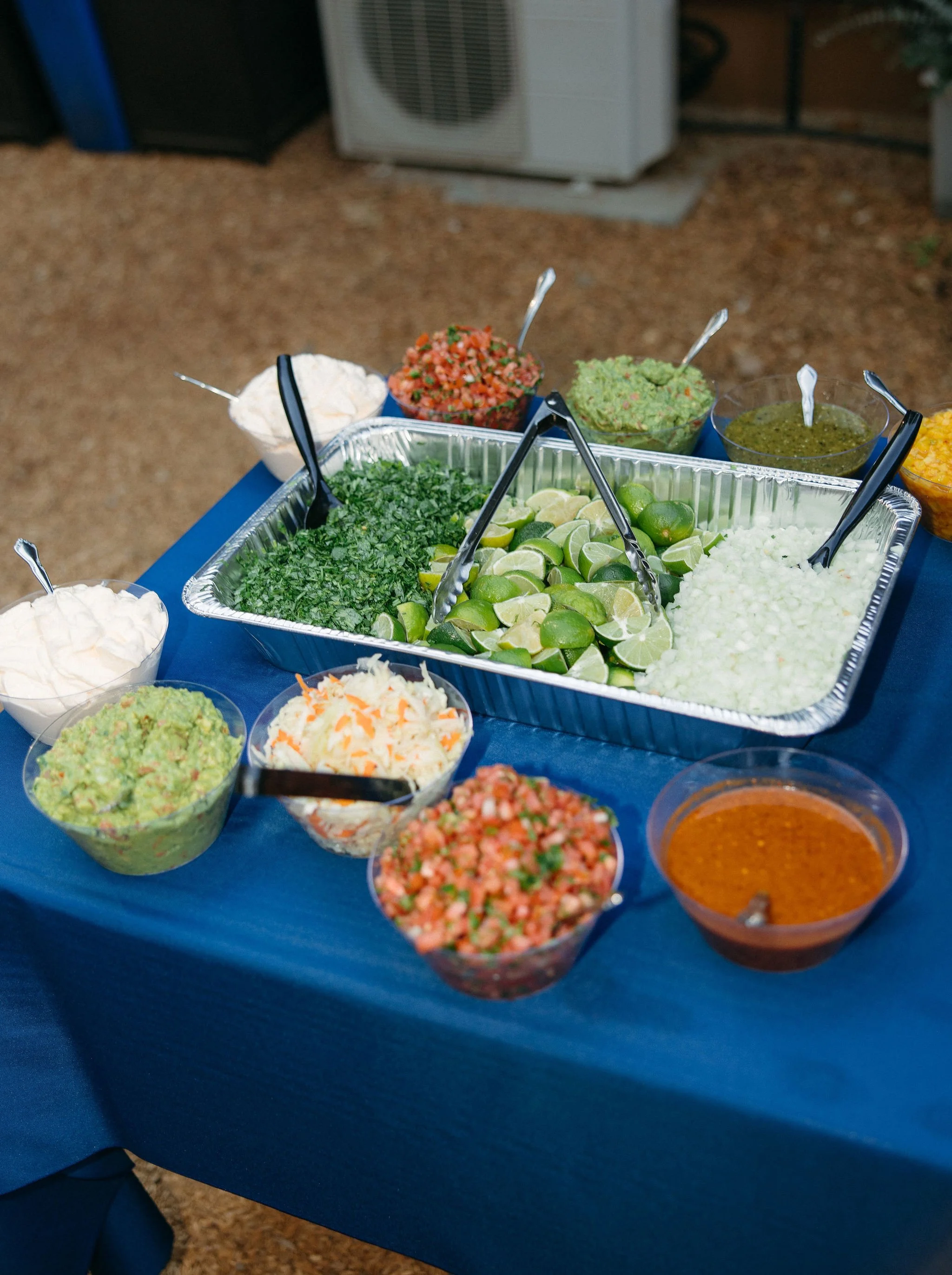 A variety of bowls containing chopped vegetables, salsa, guacamole, sour cream, shredded cheese, and lime wedges on a blue tablecloth, likely for a Mexican meal or taco bar.