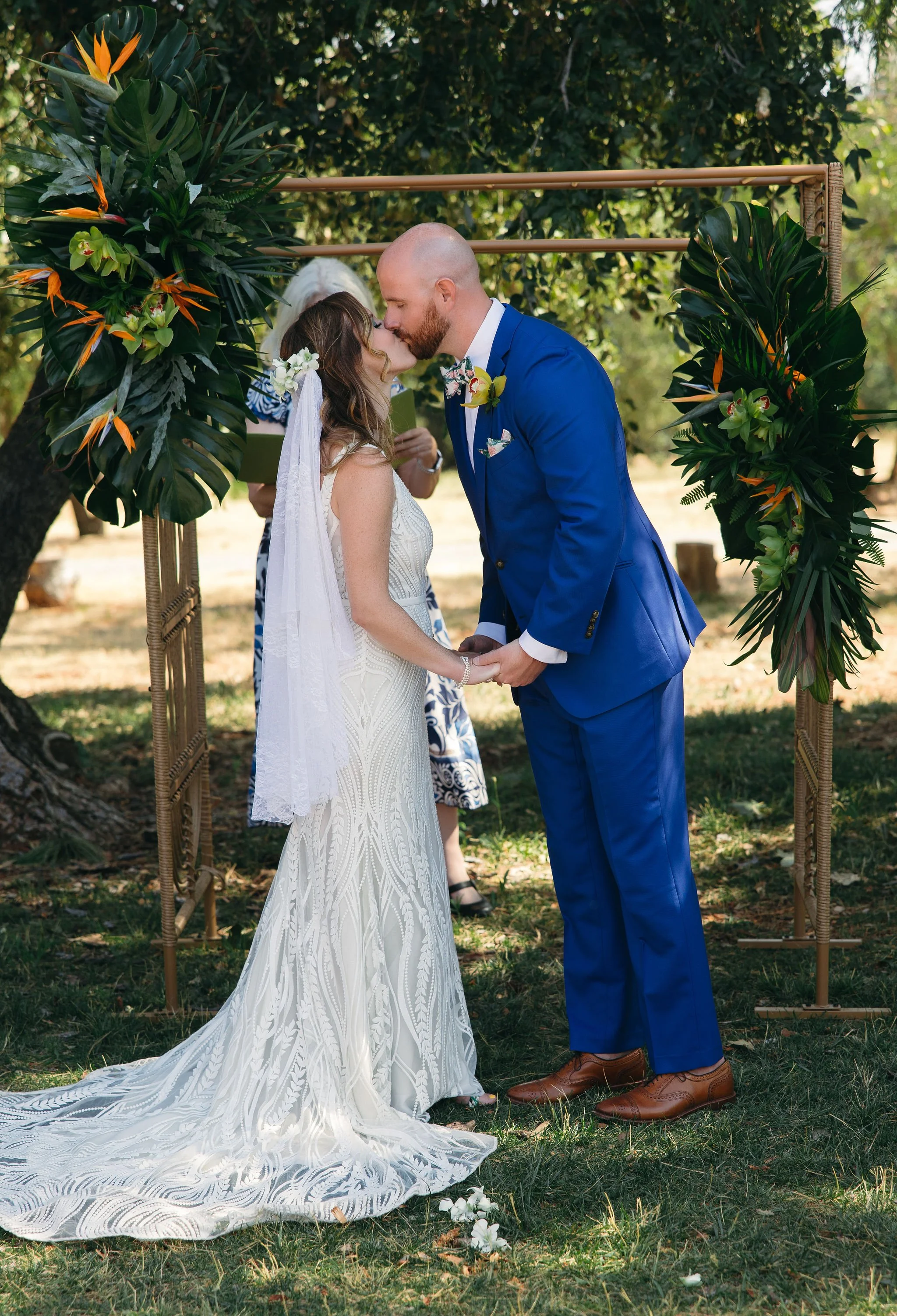 A couple kissing during their outdoor wedding ceremony under a floral arch, with an officiant in the background, surrounded by trees and grass.