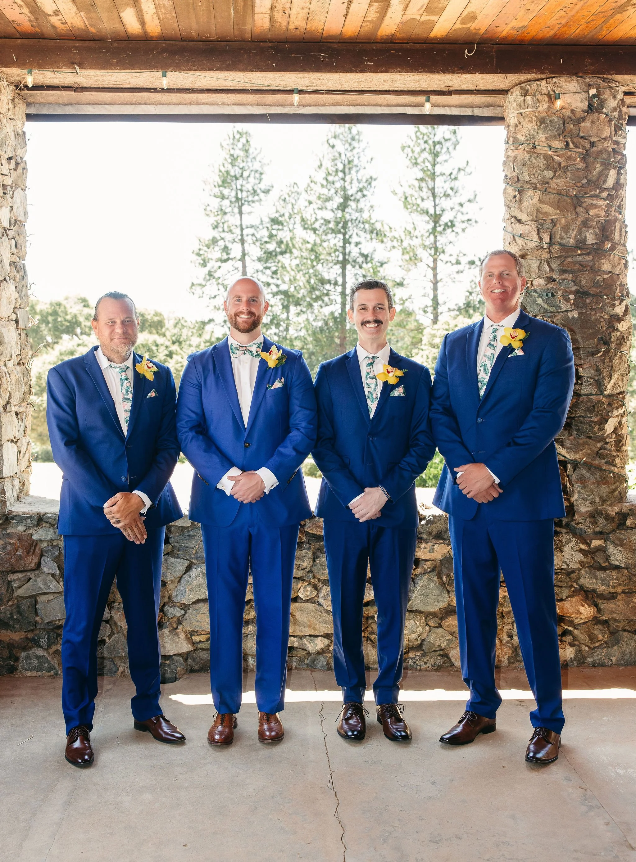 Four men in matching blue suits and floral ties, standing in front of a stone wall with a natural outdoor background, smiling.