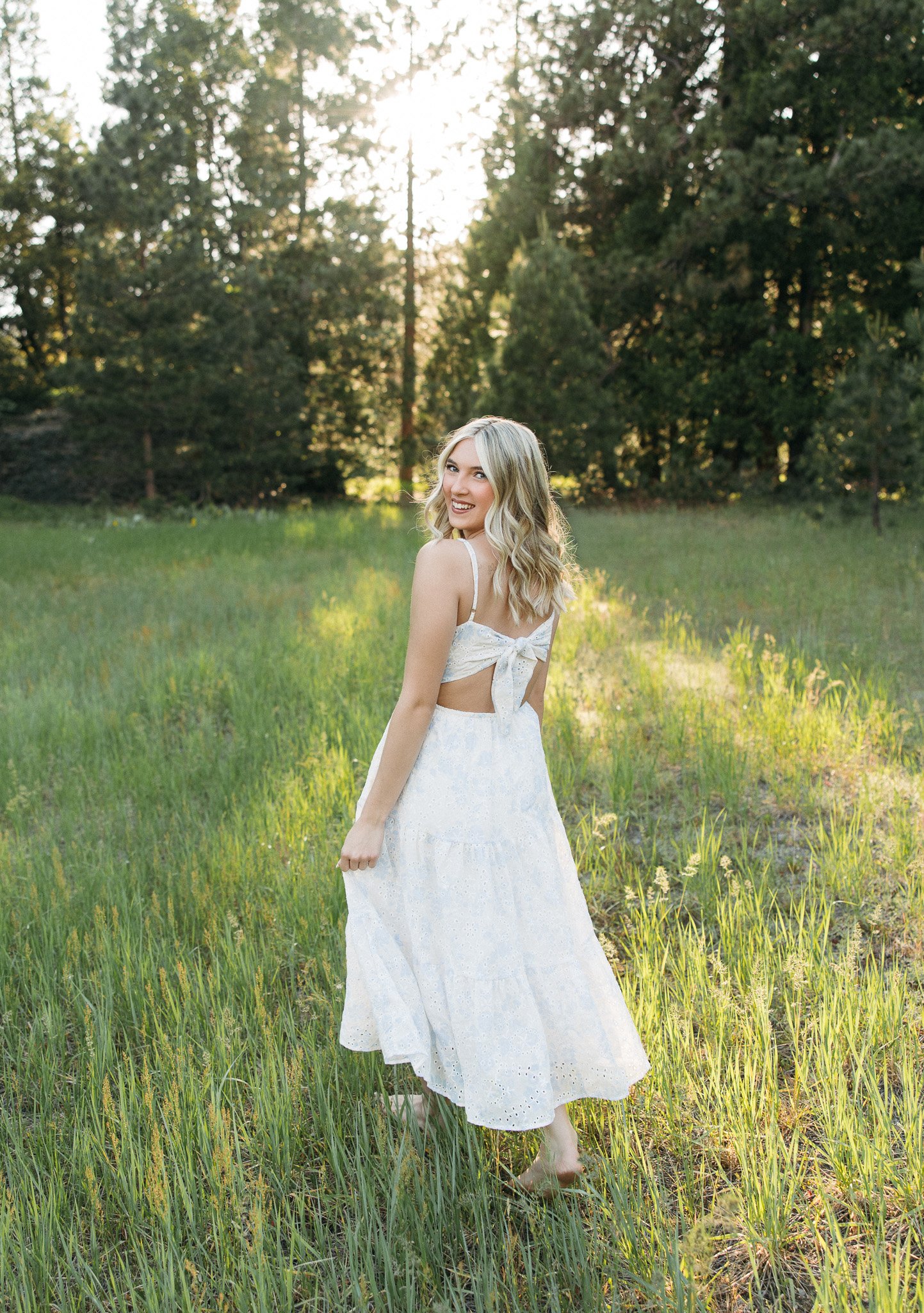 A woman with blonde hair in loose waves wearing a white summer dress, standing barefoot in a grassy field with tall grass and wildflowers, surrounded by trees, and sunlight shining through the trees in the background.