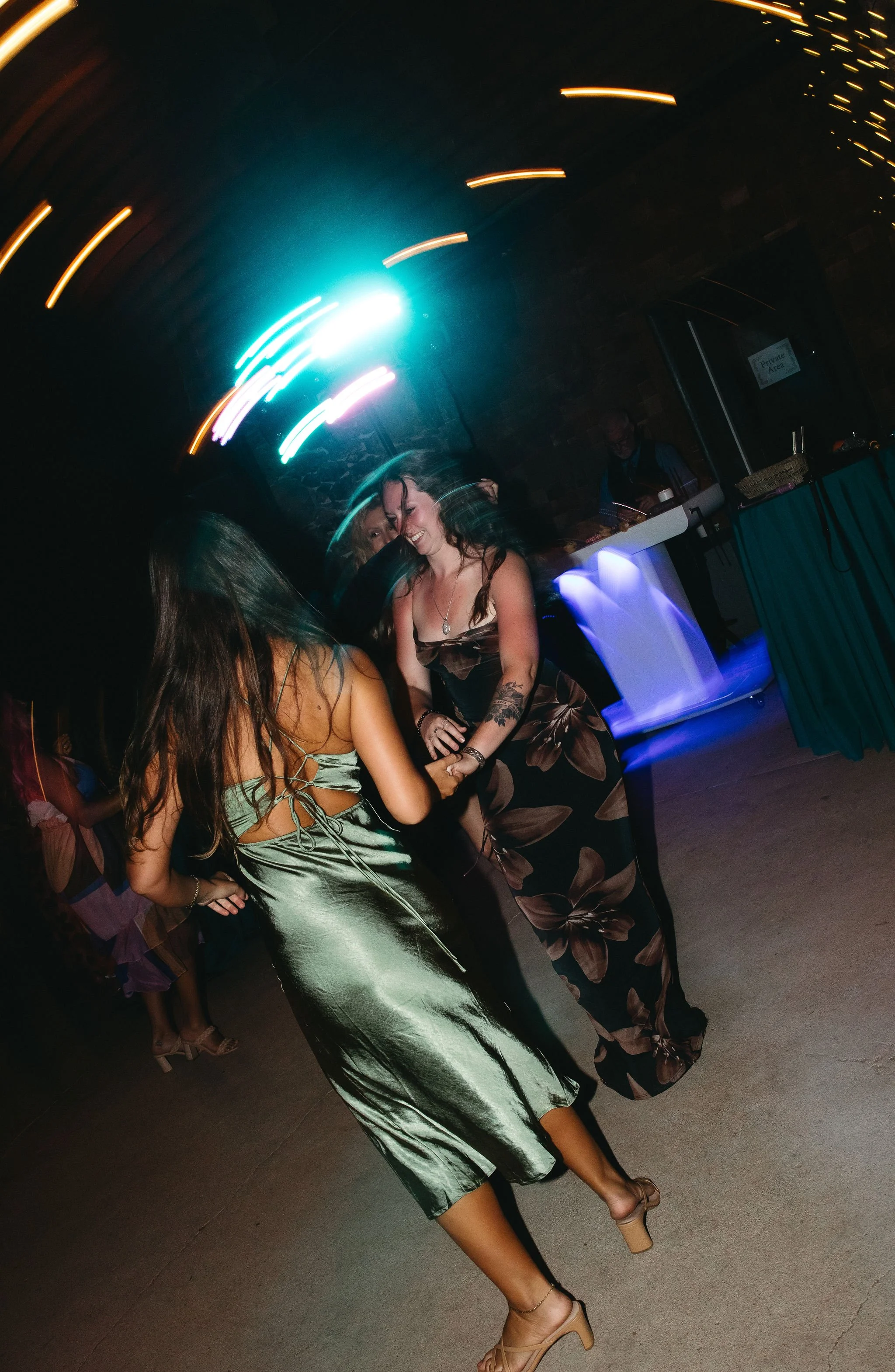 Two women dancing together at a party with colorful lights and a DJ in the background.