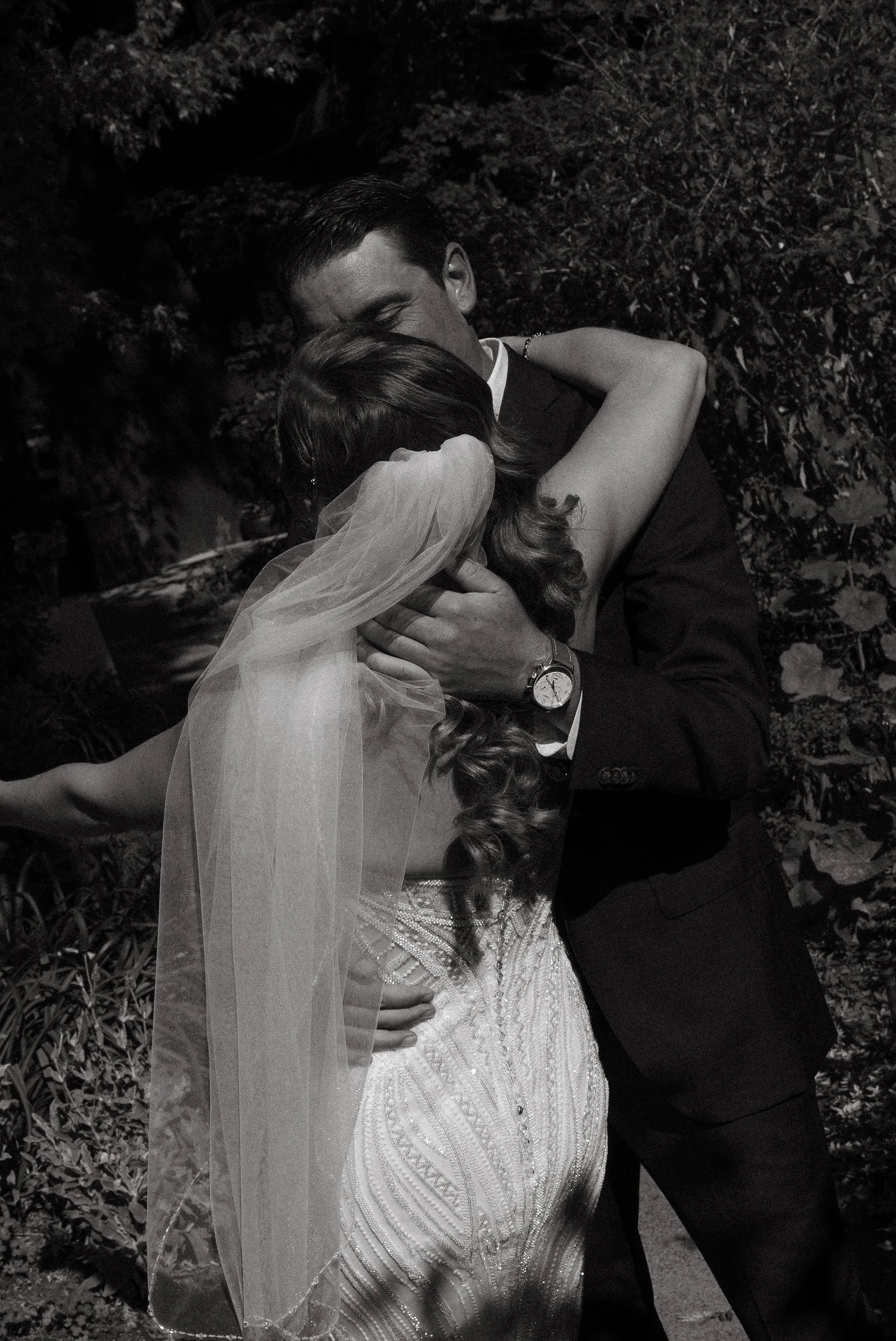 A black and white photo of a bride and groom hugging outdoors at night. The bride has a veil and a detailed dress, and the groom is in a dark suit with a watch.