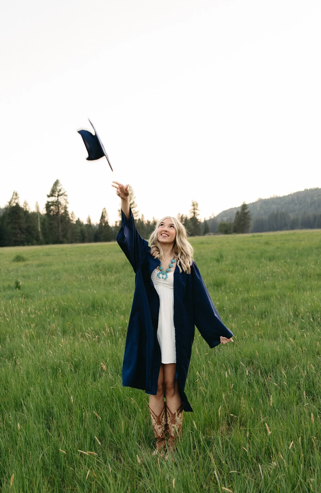 A young woman in a blue graduation gown and cowboy boots standing in a grassy field, tossing her cap in the air, with a backdrop of trees and mountains.