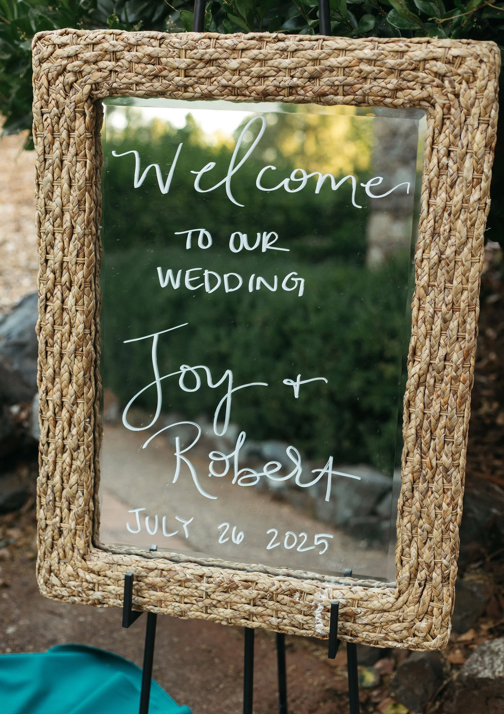 A decorated wedding welcome sign with a woven wicker frame and handwritten text on a glass panel, surrounded by greenery and rocks.