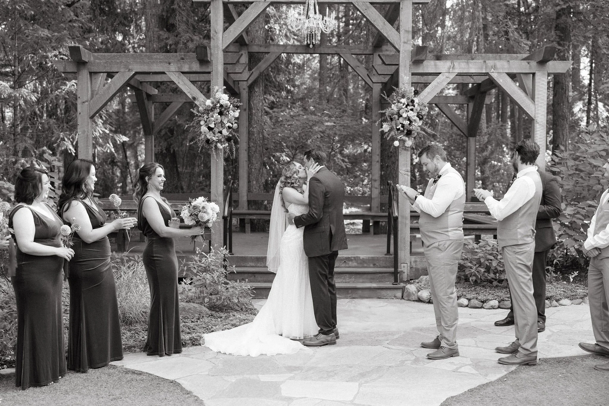 A black and white photo of a wedding ceremony outdoors, with the bride and groom kissing under a wooden arch decorated with flowers, surrounded by bridesmaids and groomsmen.