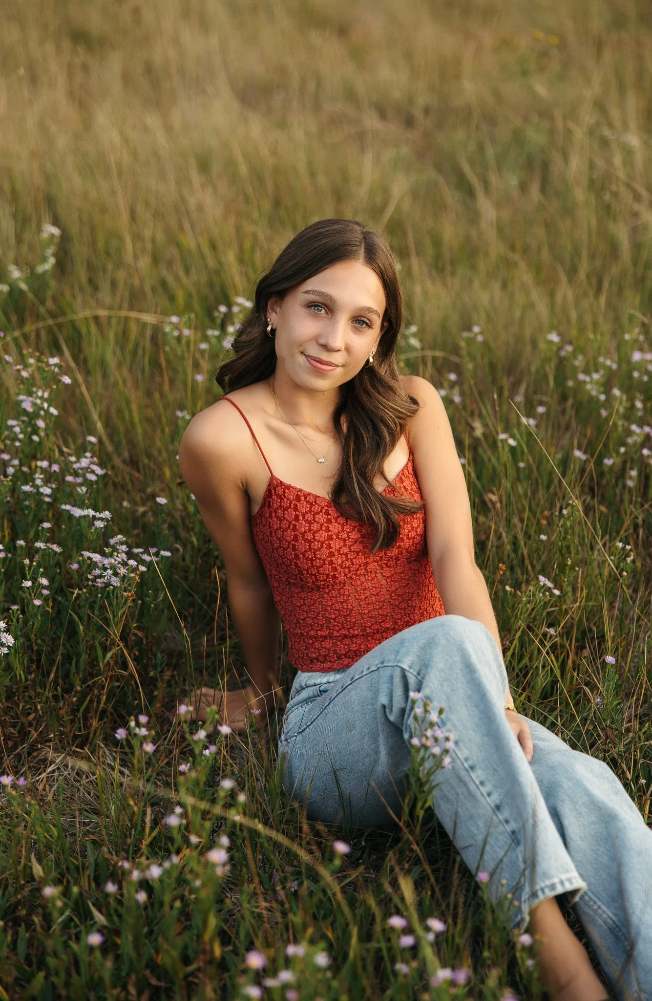 A young woman with brown hair, wearing a red lace tank top and light blue jeans, sitting in a field of tall grass and small purple flowers, looking at the camera with a slight smile.