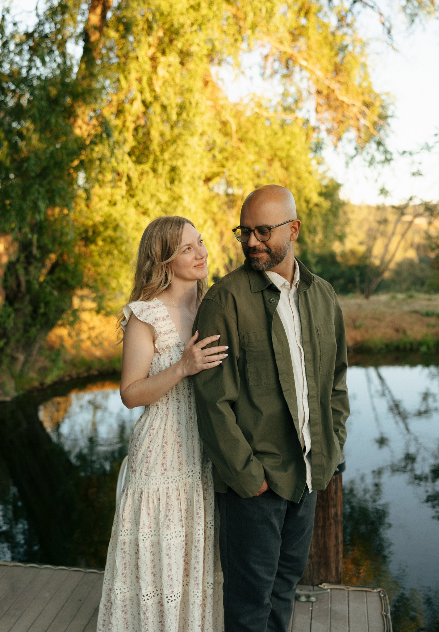 A woman with blonde wavy hair wearing a cream-colored, floral lace dress gently touching the arm of a bald man with glasses and a beard, who is wearing a green jacket and white shirt, standing outdoors near a body of water with trees in autumn foliag