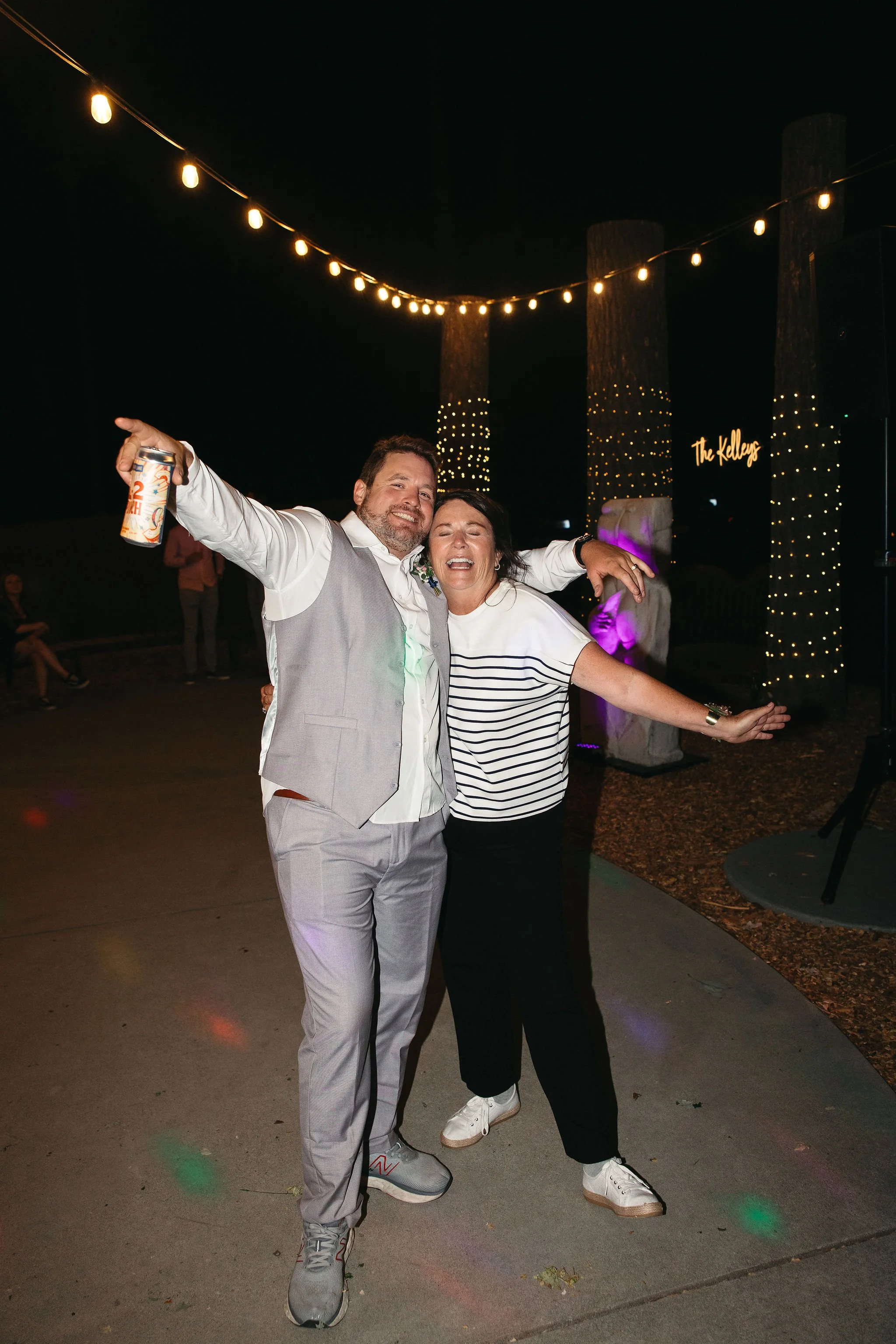 A man and woman are dancing and enjoying themselves at an outdoor night event, with string lights hanging above and a sign that says 'The Kellys' in the background.