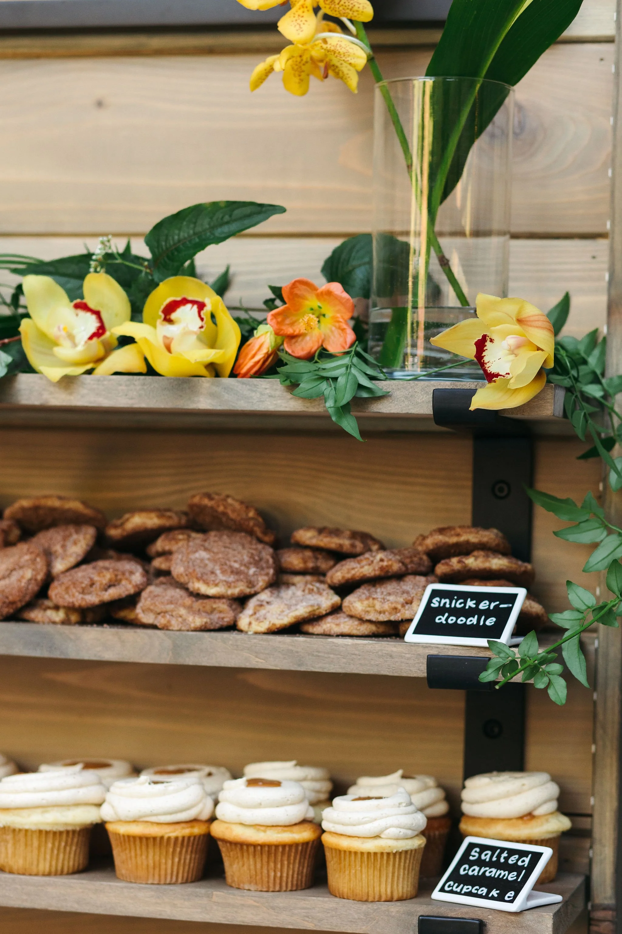 Display of baked goods on wooden shelves including cookies, cupcakes with white frosting and caramel drizzle, and decorative flowers in a glass vase.