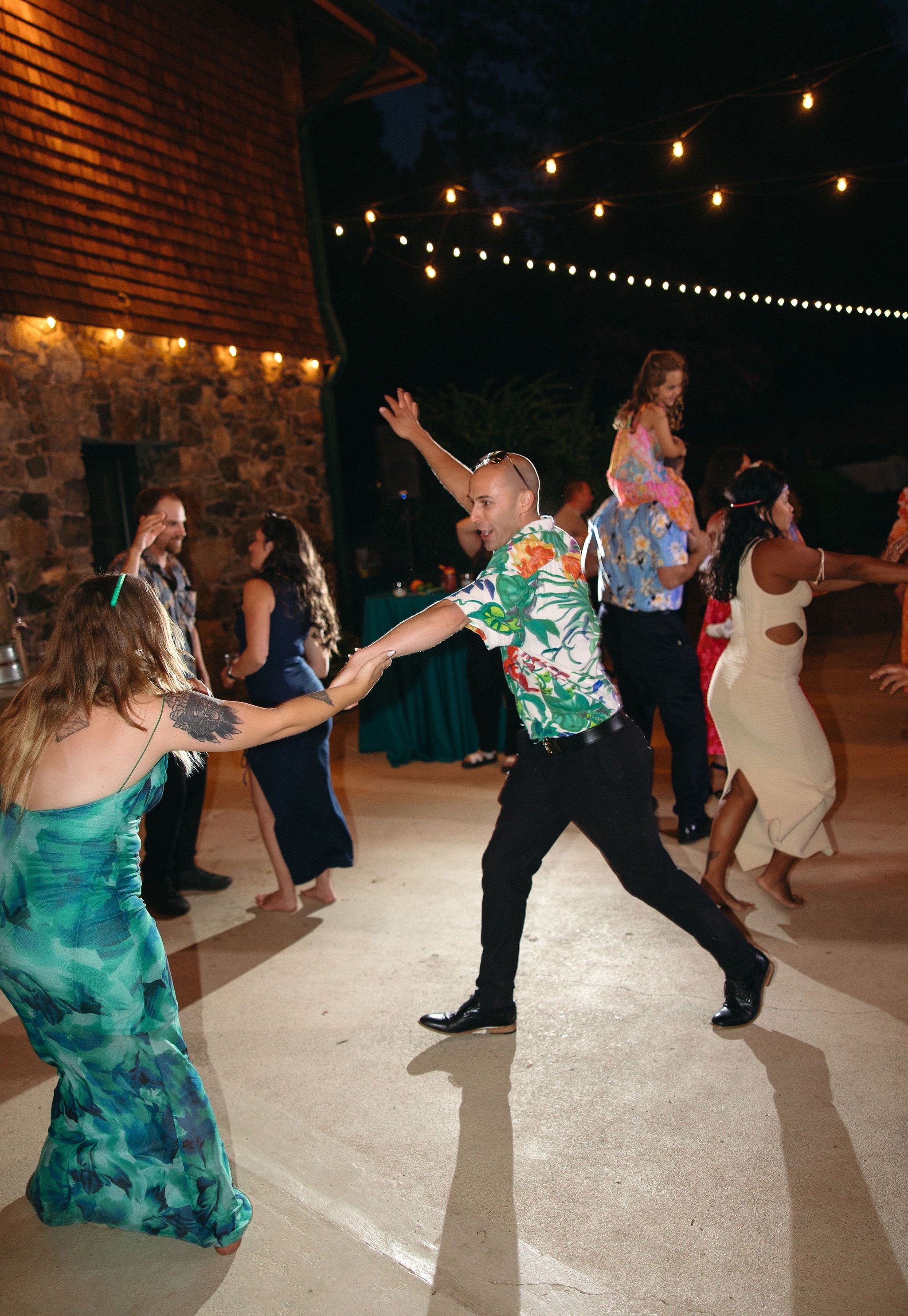 People dancing at an outdoor party during the night under string lights, with a stone wall in the background.