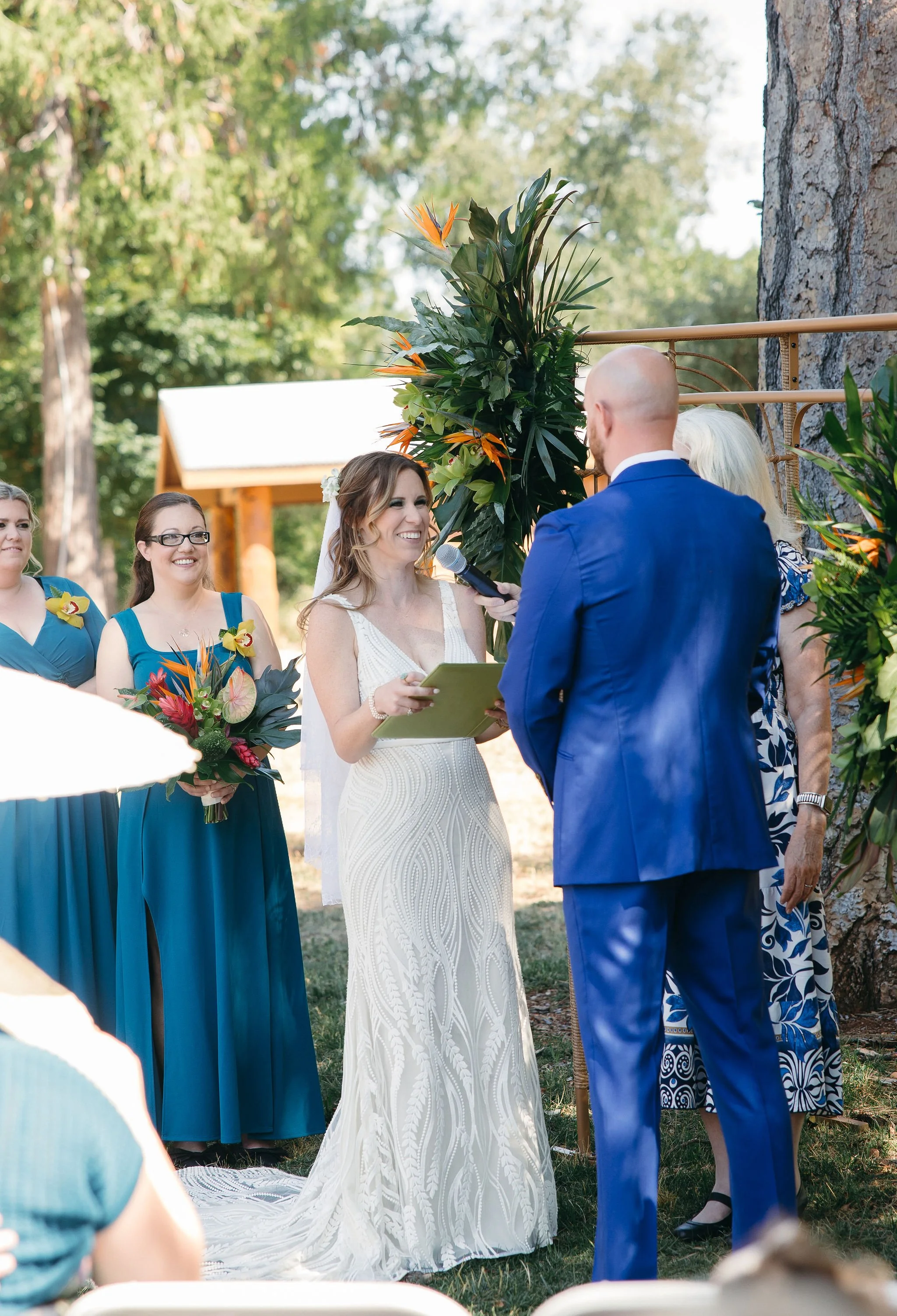 A bride in a white dress is smiling during her wedding ceremony outdoors, standing next to a man in a blue suit. An officiant holds a microphone to the bride. Two bridesmaids in blue dresses holding bouquets stand nearby, with trees and a small woode