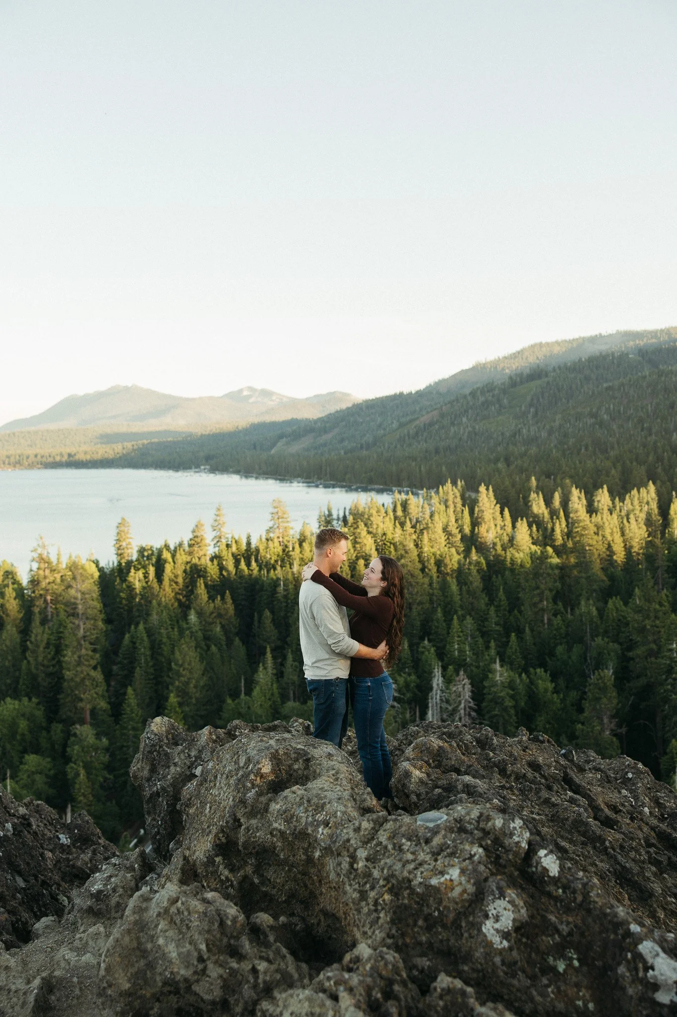 A couple stands on large rocks surrounded by a forest of pine trees, with mountains and a lake in the background, during the evening.