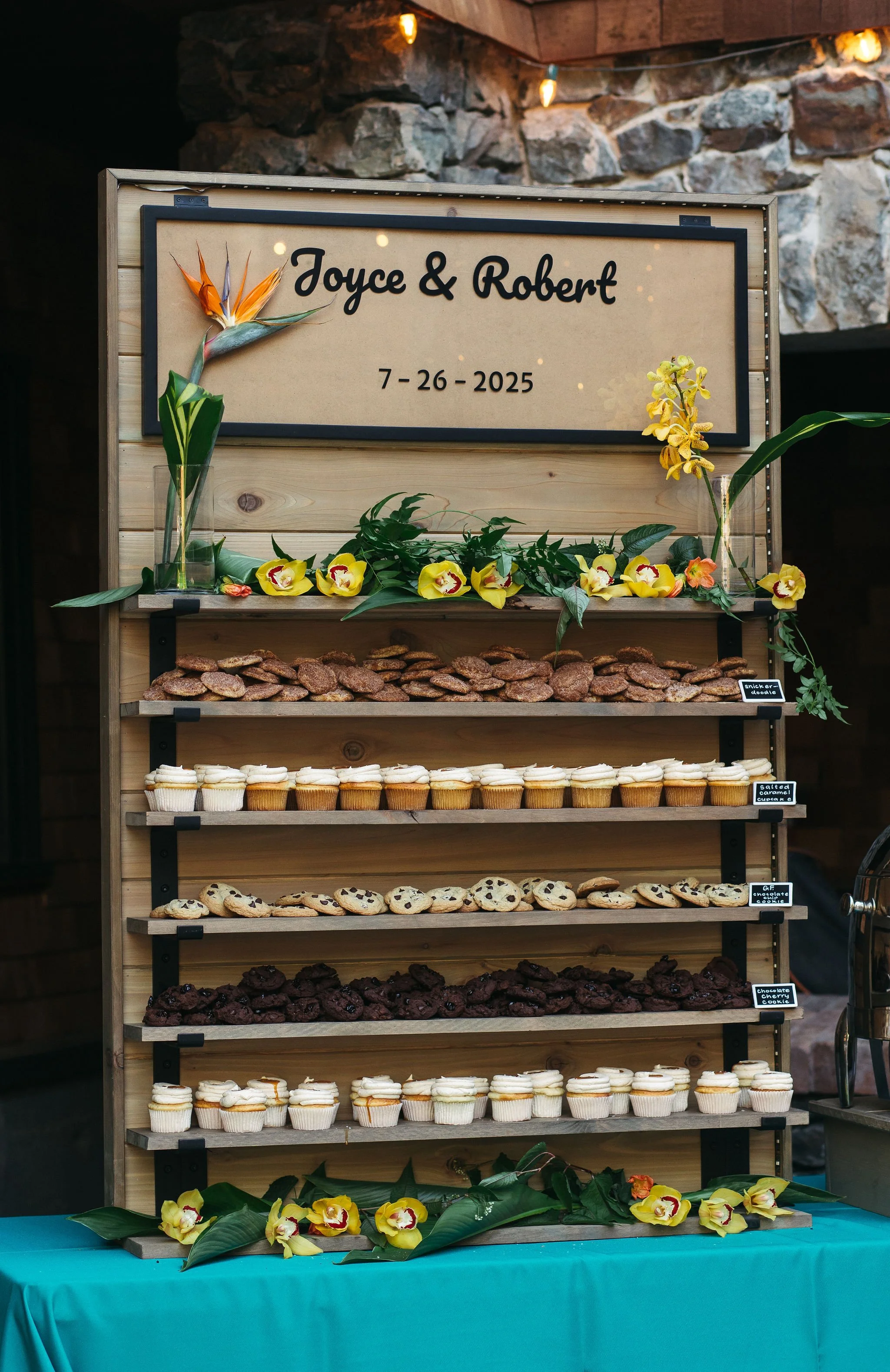 A dessert display with cupcakes, cookies, and chocolate chip cookies, decorated with yellow orchids and green leaves, with a sign reading 'Joyce & Robert' and date '7-26-2025'.