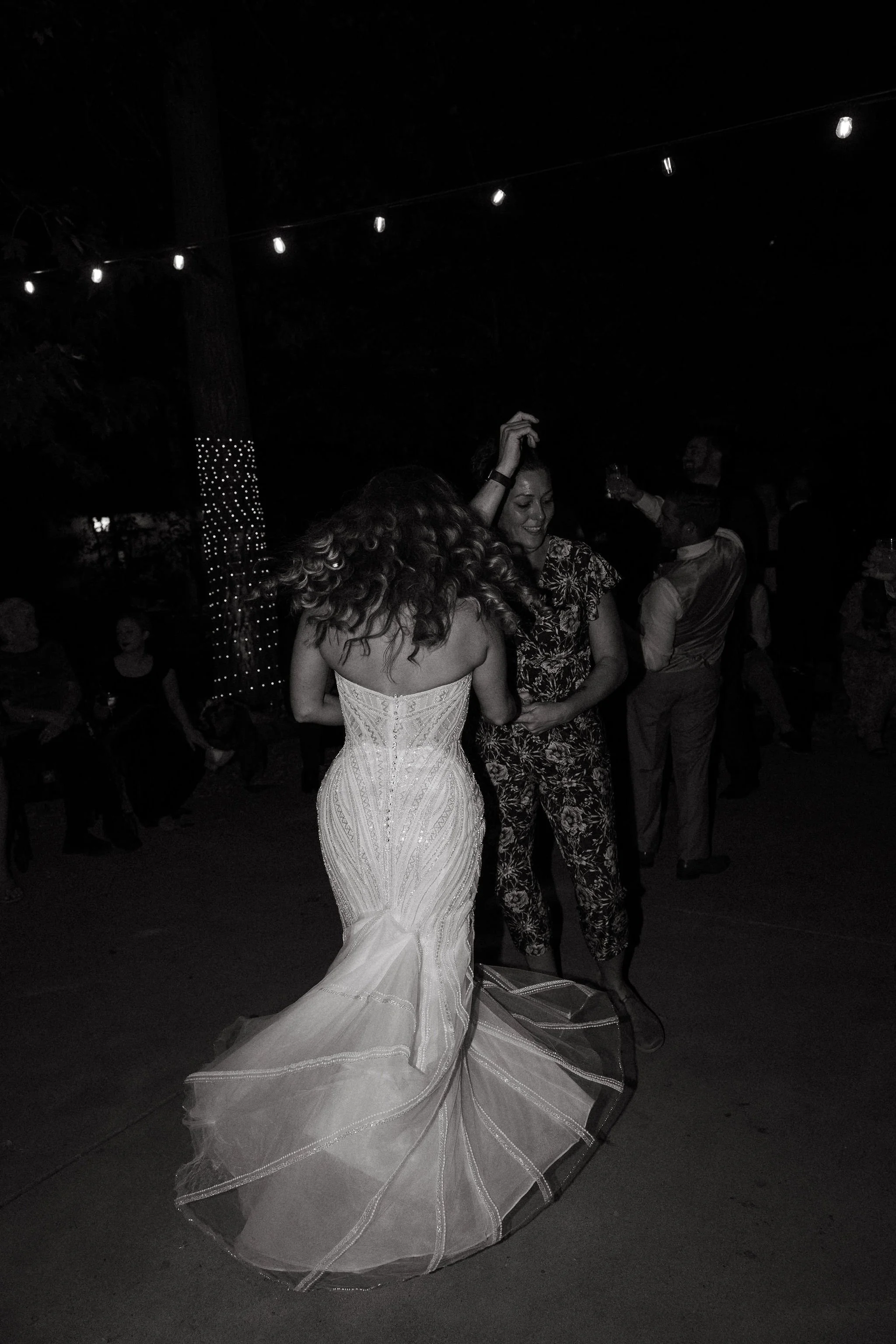 Women dancing at a nighttime celebration, one in a wedding dress, others in casual attire, outdoor setting with string lights.
