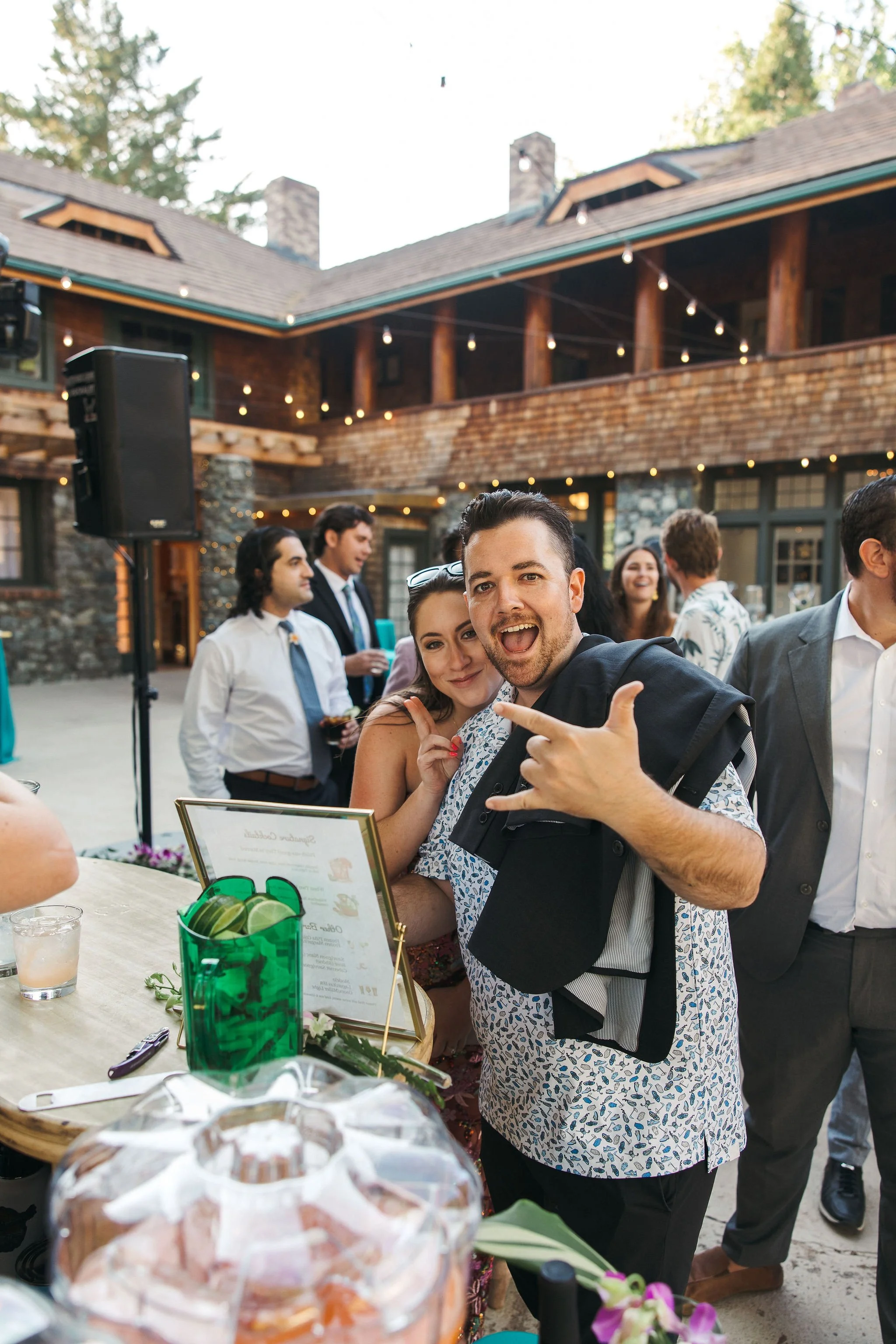 People enjoying a celebration outdoors near a rustic building with string lights, with a man making a rock and roll hand gesture and a woman smiling next to him.