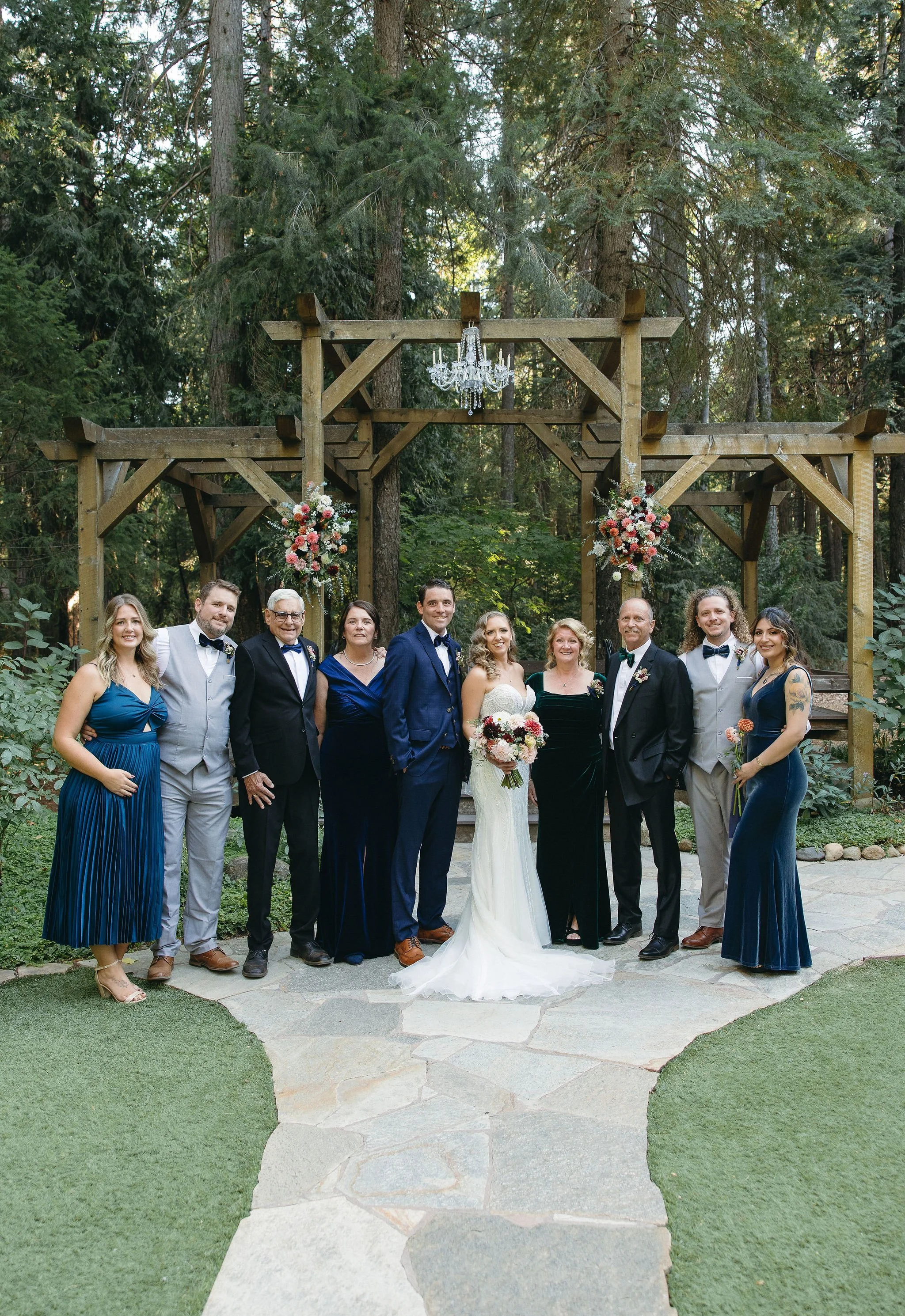 A wedding party with nine people, including a bride in a white gown holding a bouquet, and a groom in a blue suit, standing outdoors on a stone path in front of a wooden arbor decorated with pink and white flowers and a chandelier, surrounded by tall
