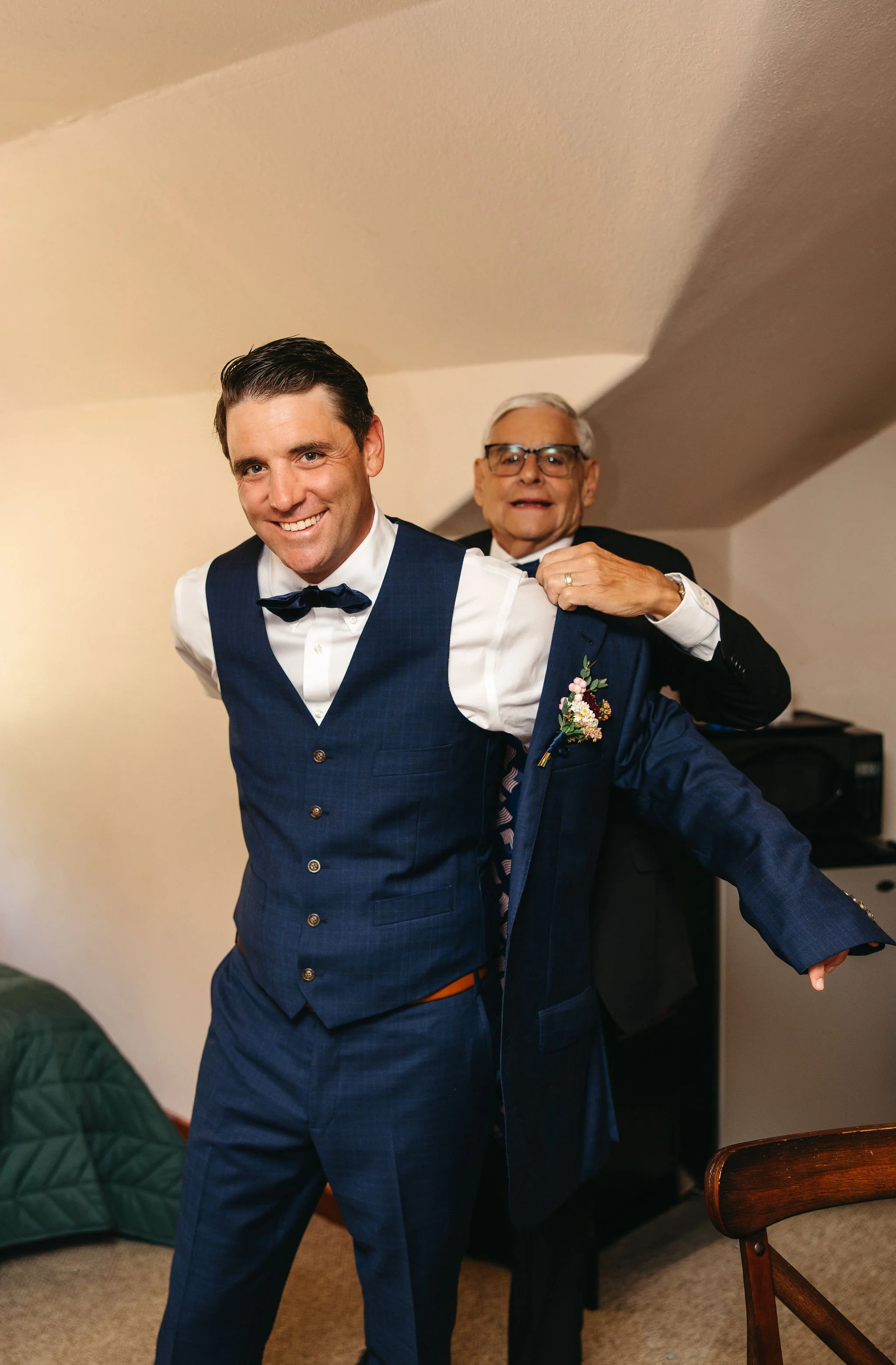 A happy man in a blue suit and bow tie having his jacket put on by an older man in a black suit, in a cozy room with a beige wall and wooden chair.