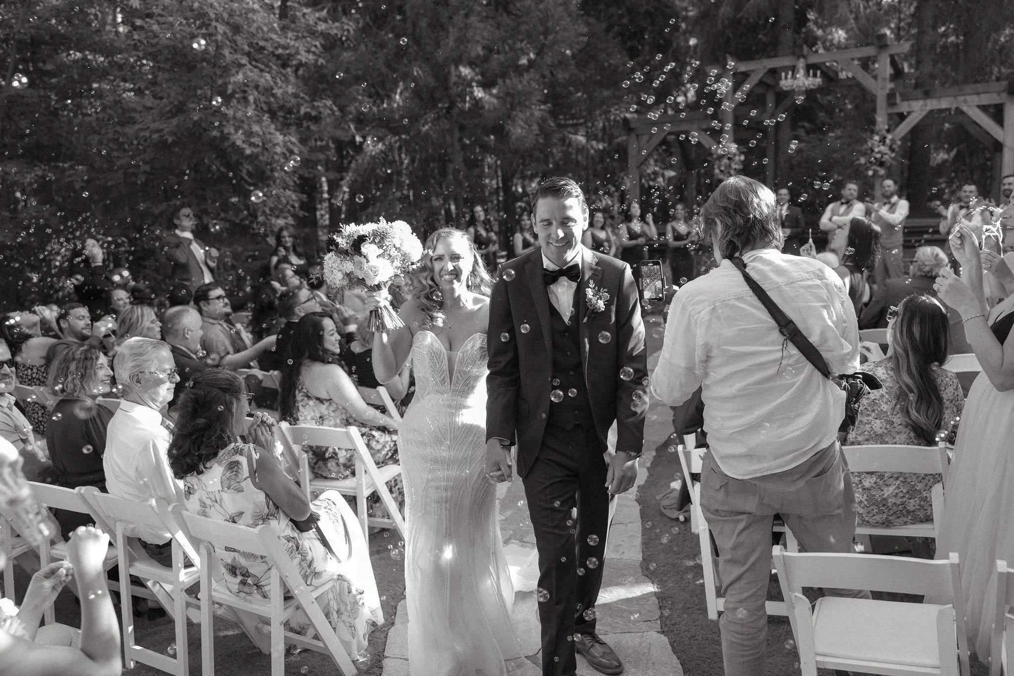 Black and white photo of a newlywed couple walking down the aisle at their outdoor wedding, surrounded by seated guests and bubbles in the air.