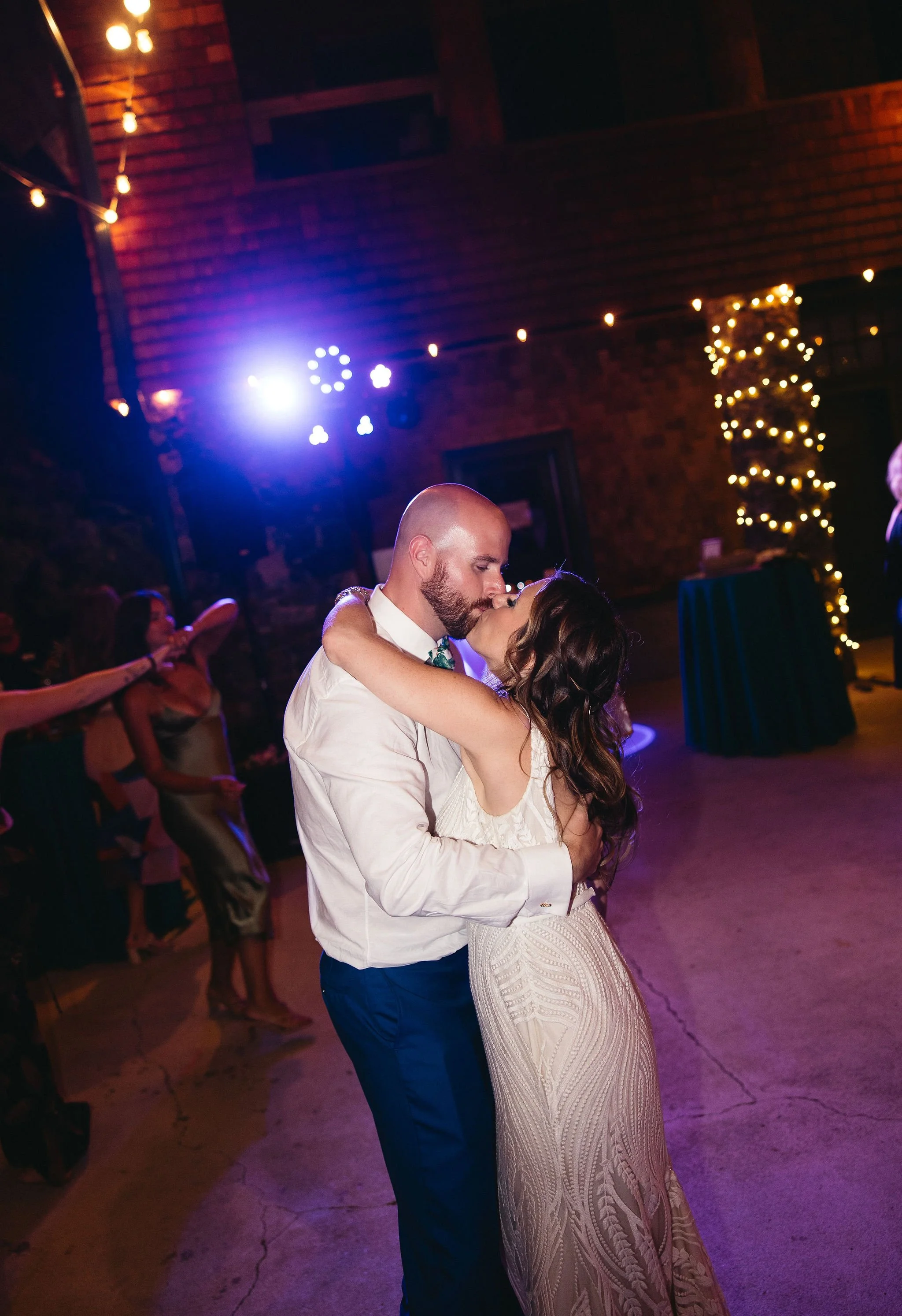 A couple sharing a kiss during their wedding reception dance, surrounded by guests in a warmly lit venue with string lights and a decorated pillar.