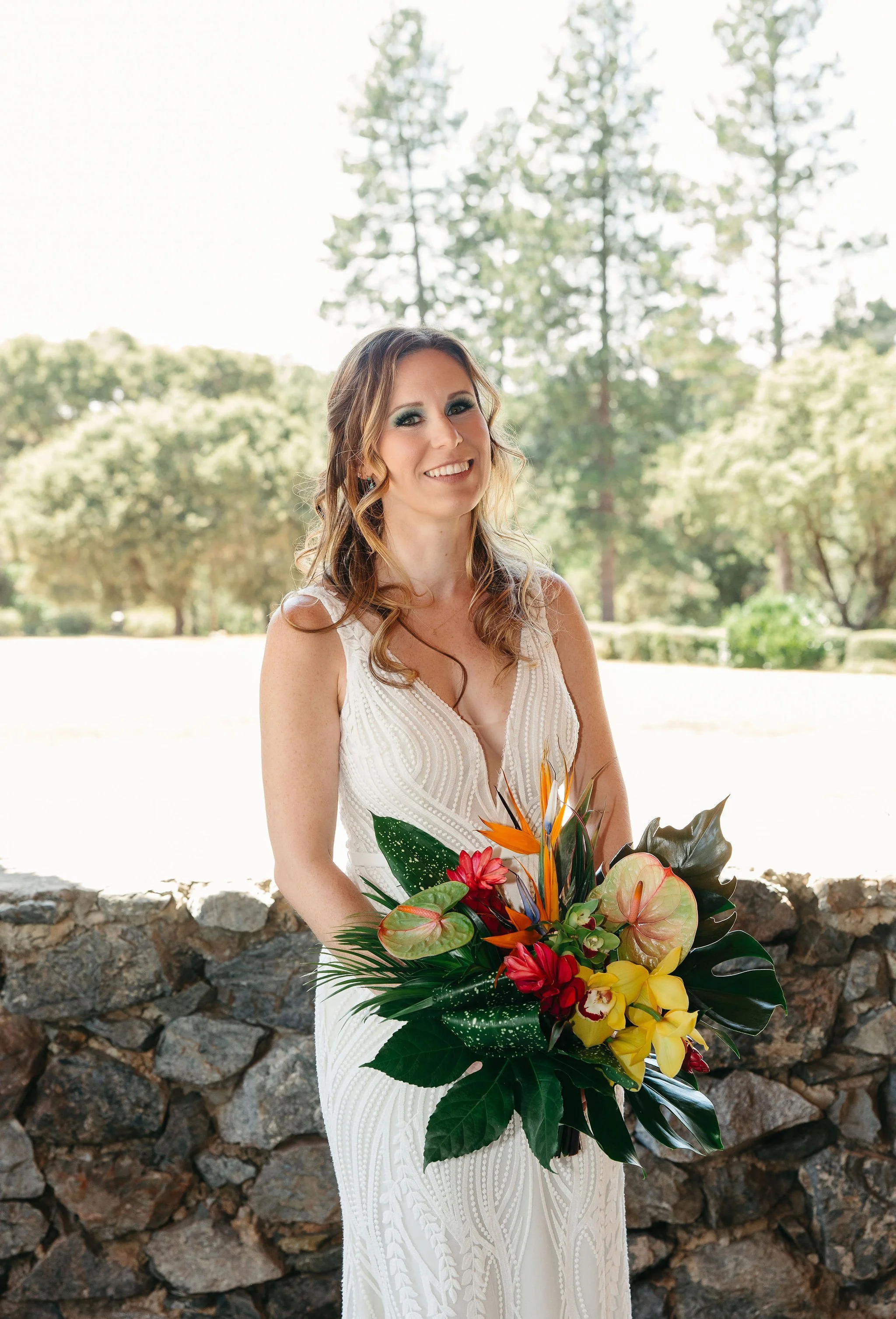 Bride in a white wedding dress holding a colorful tropical bouquet, standing outdoors against a stone wall with green trees in the background.