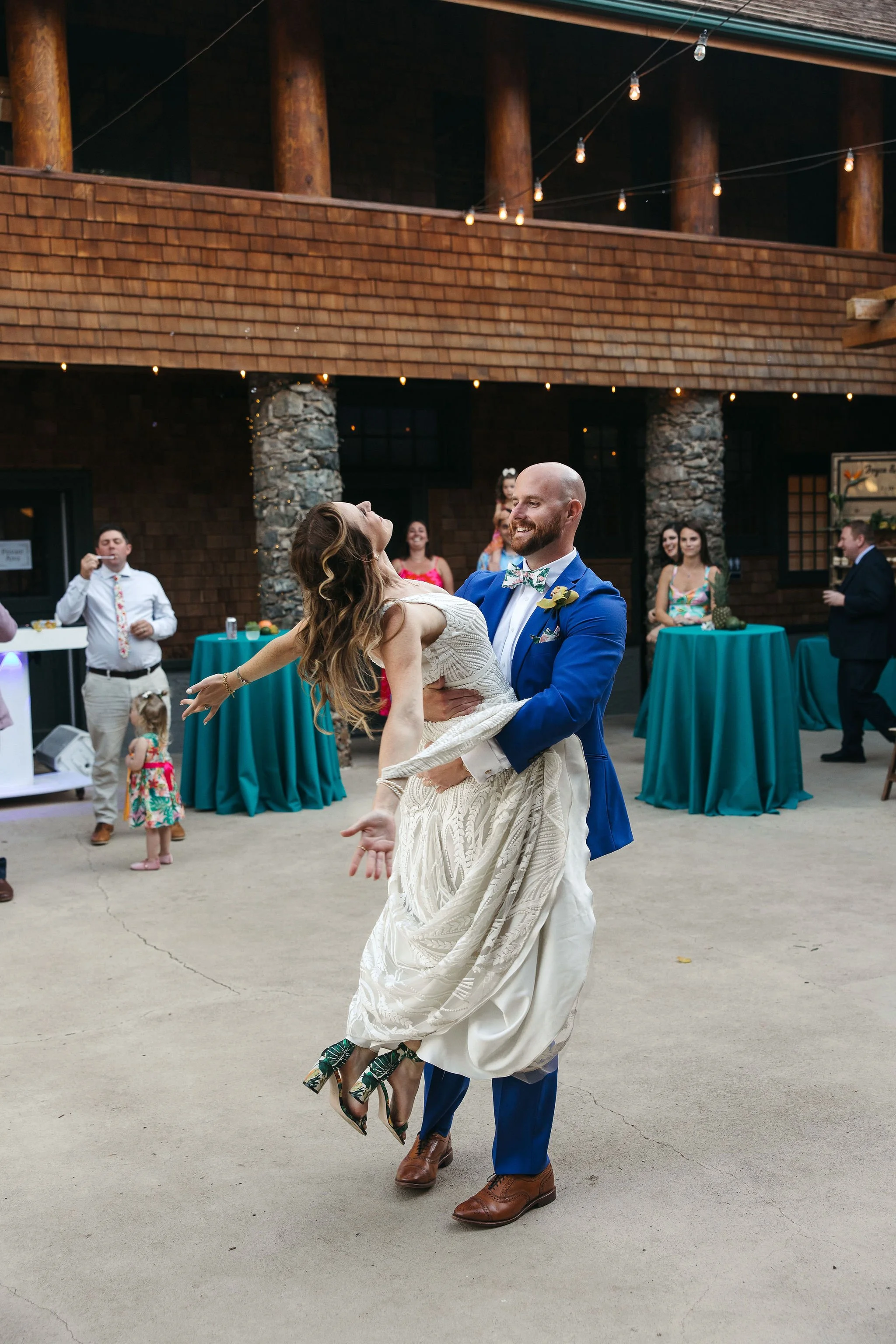 A man in a blue suit and bow tie is dancing with a woman in a wedding dress at an outdoor wedding reception, with guests and string lights in the background.
