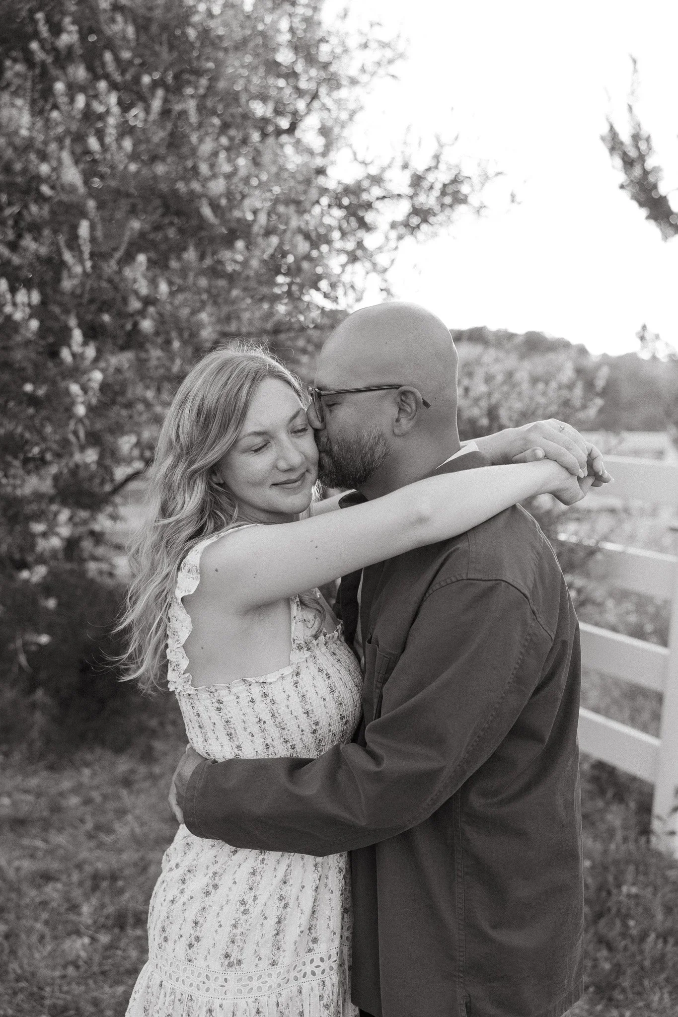 A black-and-white photo of a man and woman embracing outdoors. The woman is smiling with her eyes closed, while the man is kissing her forehead. The background features trees and a white fence.