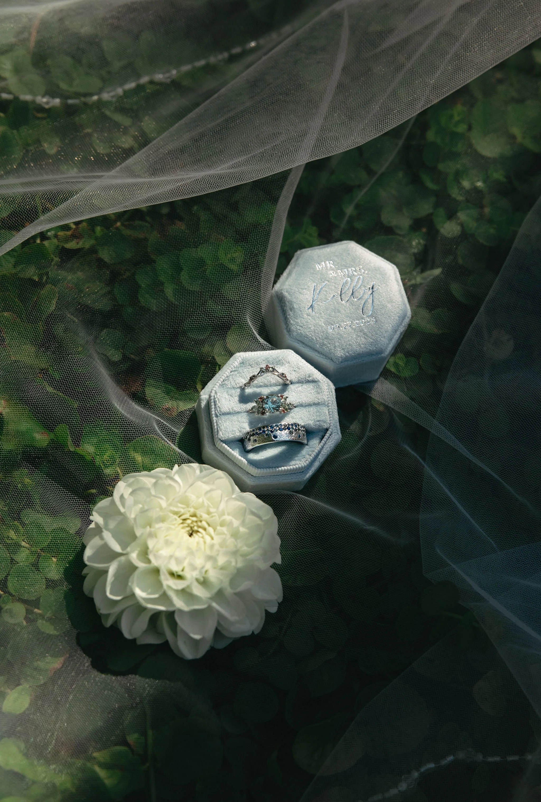 A white flower next to a jewelry box with rings inside, and another jewelry box labeled 'Mr. and Mrs. Kelly' on a green leafy background covered with sheer fabric.