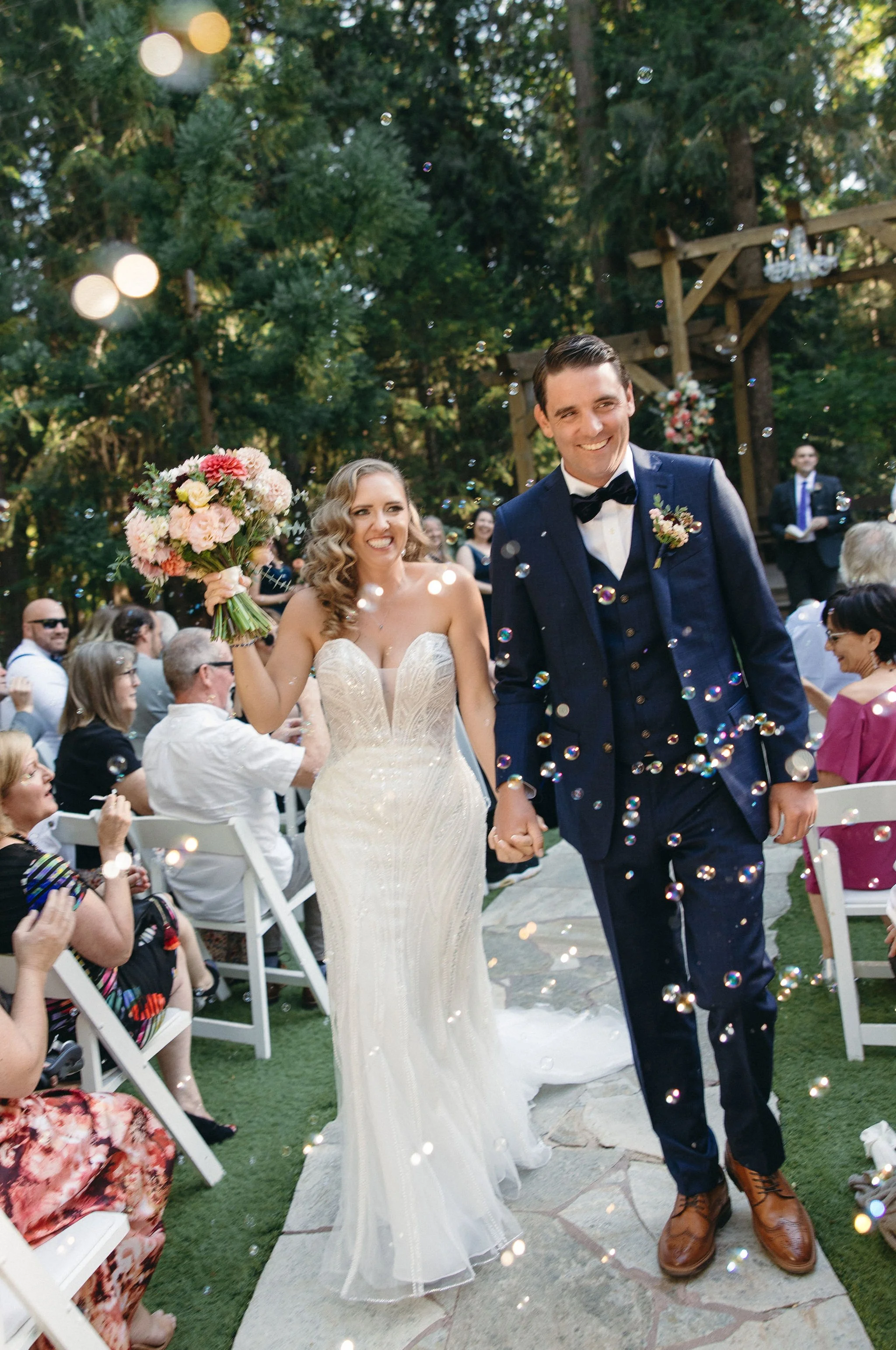 Bride and groom walking down the aisle at their outdoor wedding, holding hands, surrounded by guests, with bubbles floating in the air.